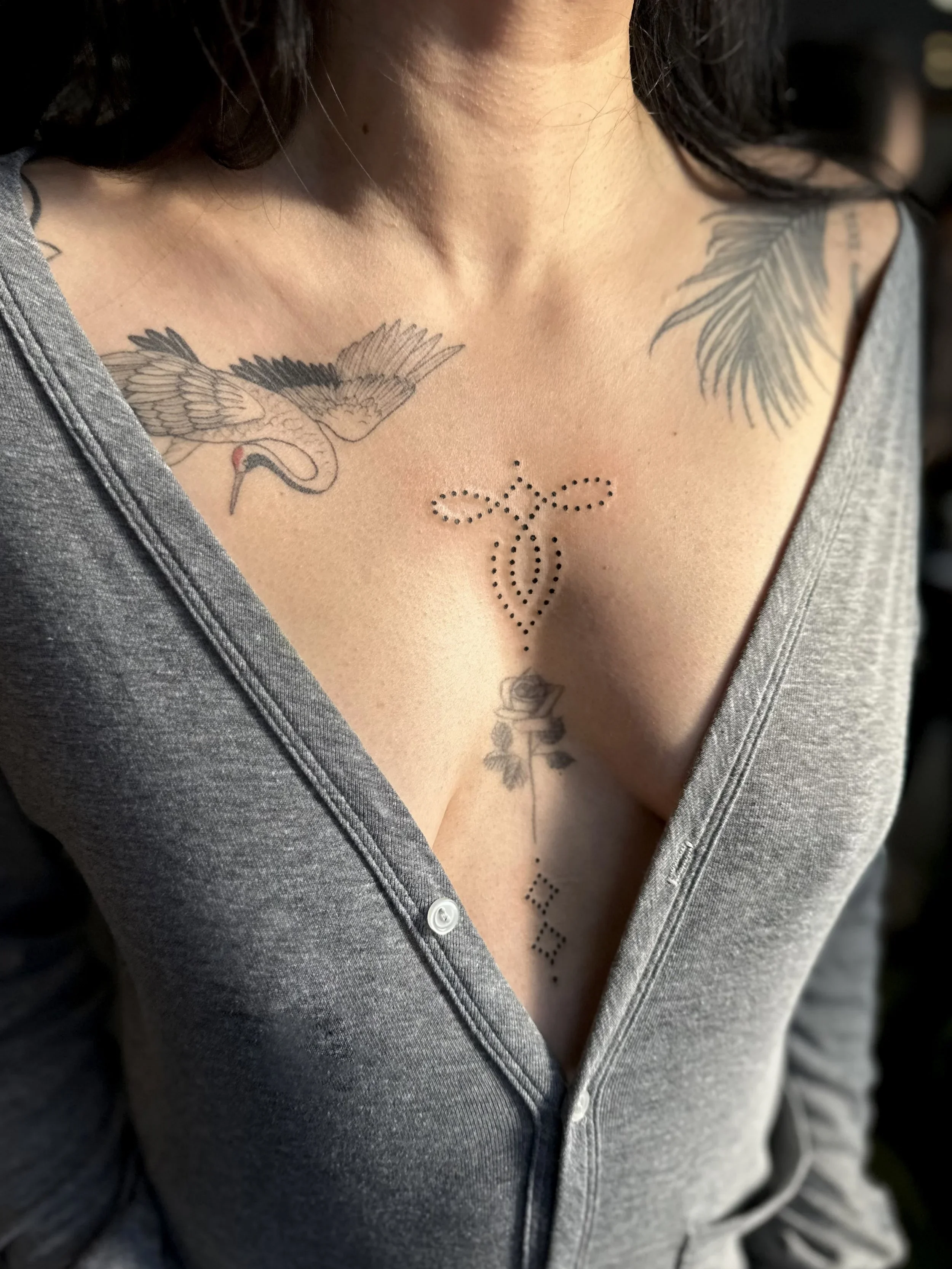 Close-up of a woman's chest showing tattoos of a bird and a heart with dotted lines on her skin. She is wearing a gray button-up shirt partially open.