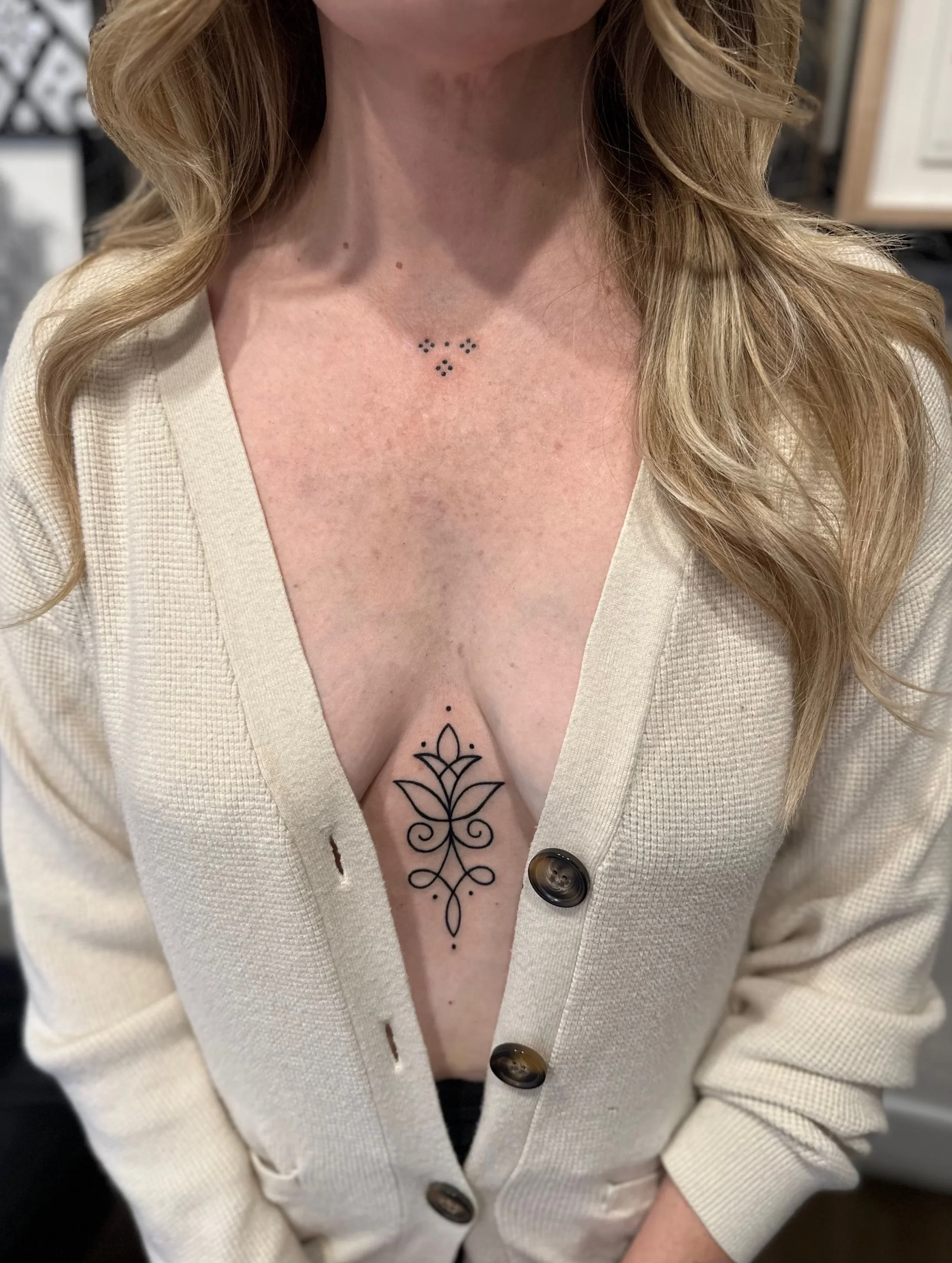 Close-up of a woman's chest with a black ink floral tattoo with symmetrical patterns and dots on her sternum, wearing a cream-colored button-up shirt.