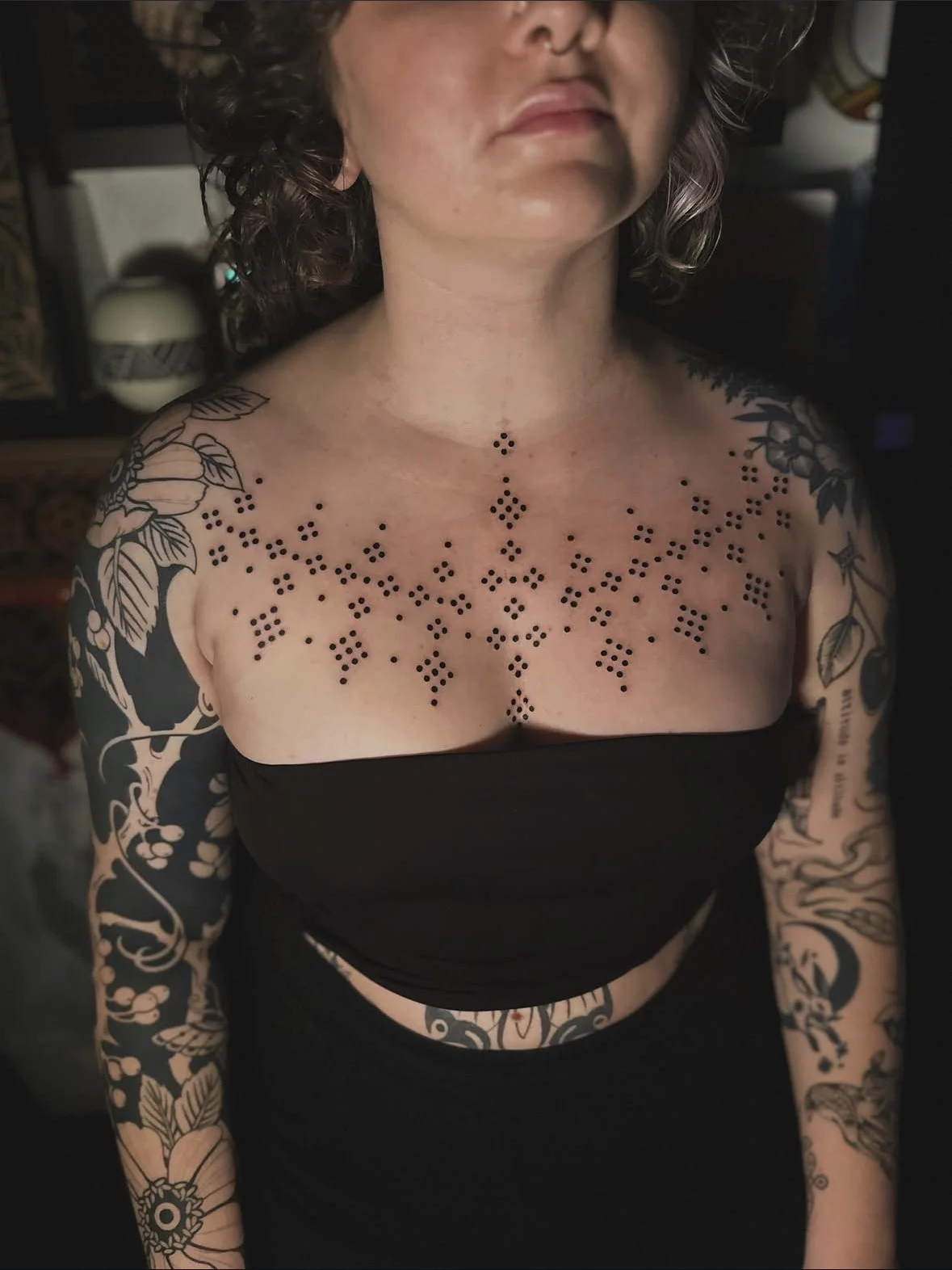 A woman with shoulder-length curly hair wearing a black strapless top and tattoos on her shoulders, chest, and arms. The tattoos are floral and ornamental, with a dotwork pattern on her chest and collarbone area.