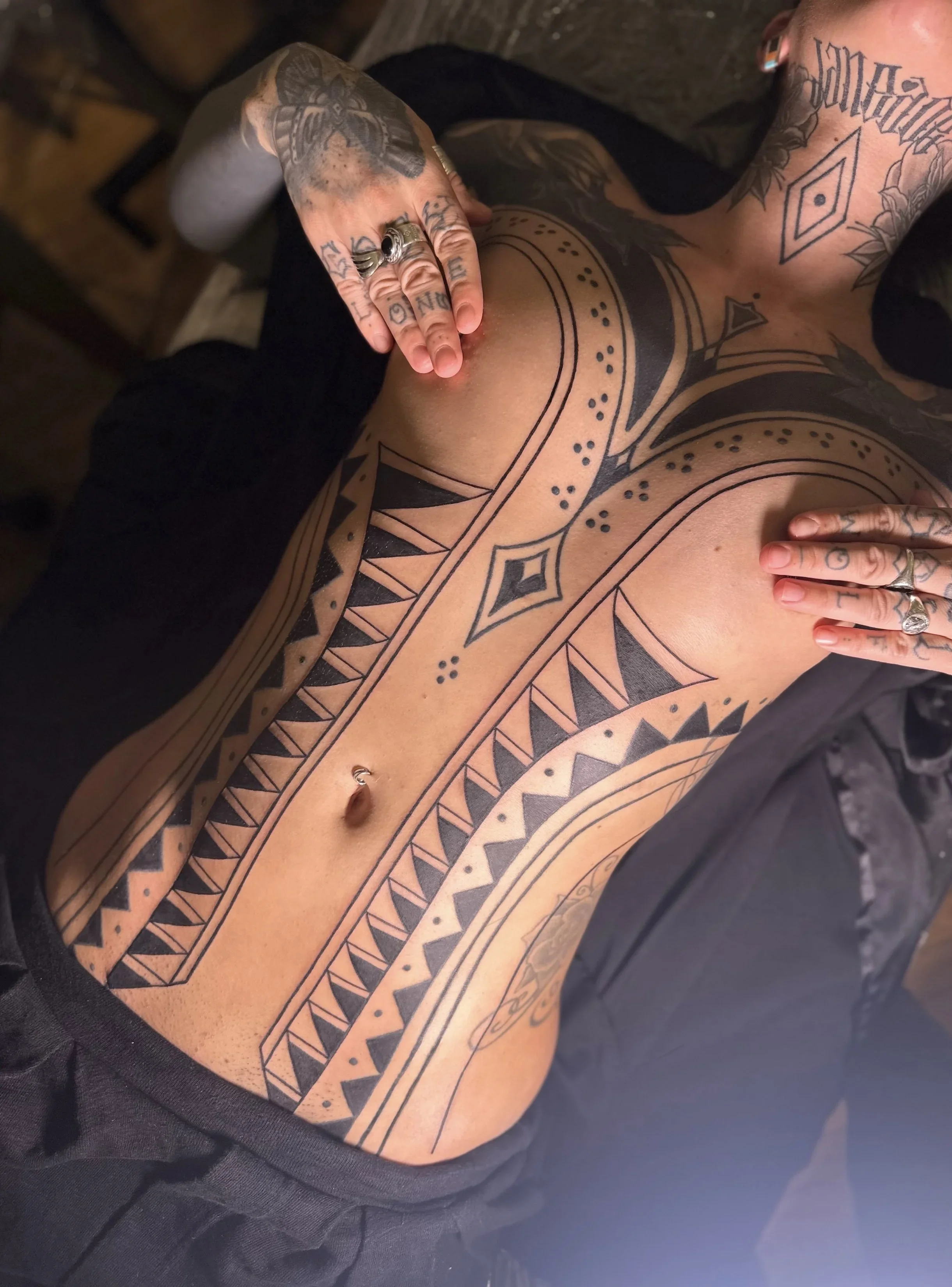 A person with tattoos on their chest and arm is resting on a black bag, with a wooden floor in the background.