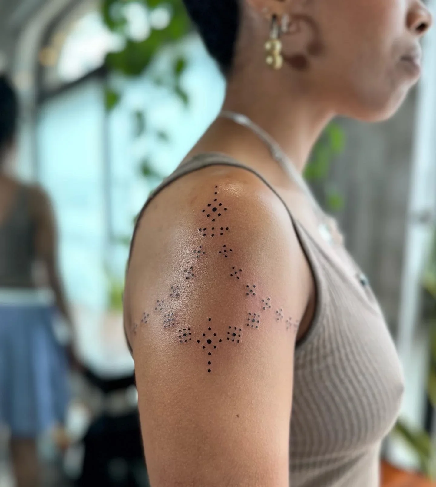 Close-up of a woman's upper arm with a tattoo of a constellation made of small dots arranged in a pattern. The woman is wearing a sleeveless top and has short hair, with earrings and necklace.