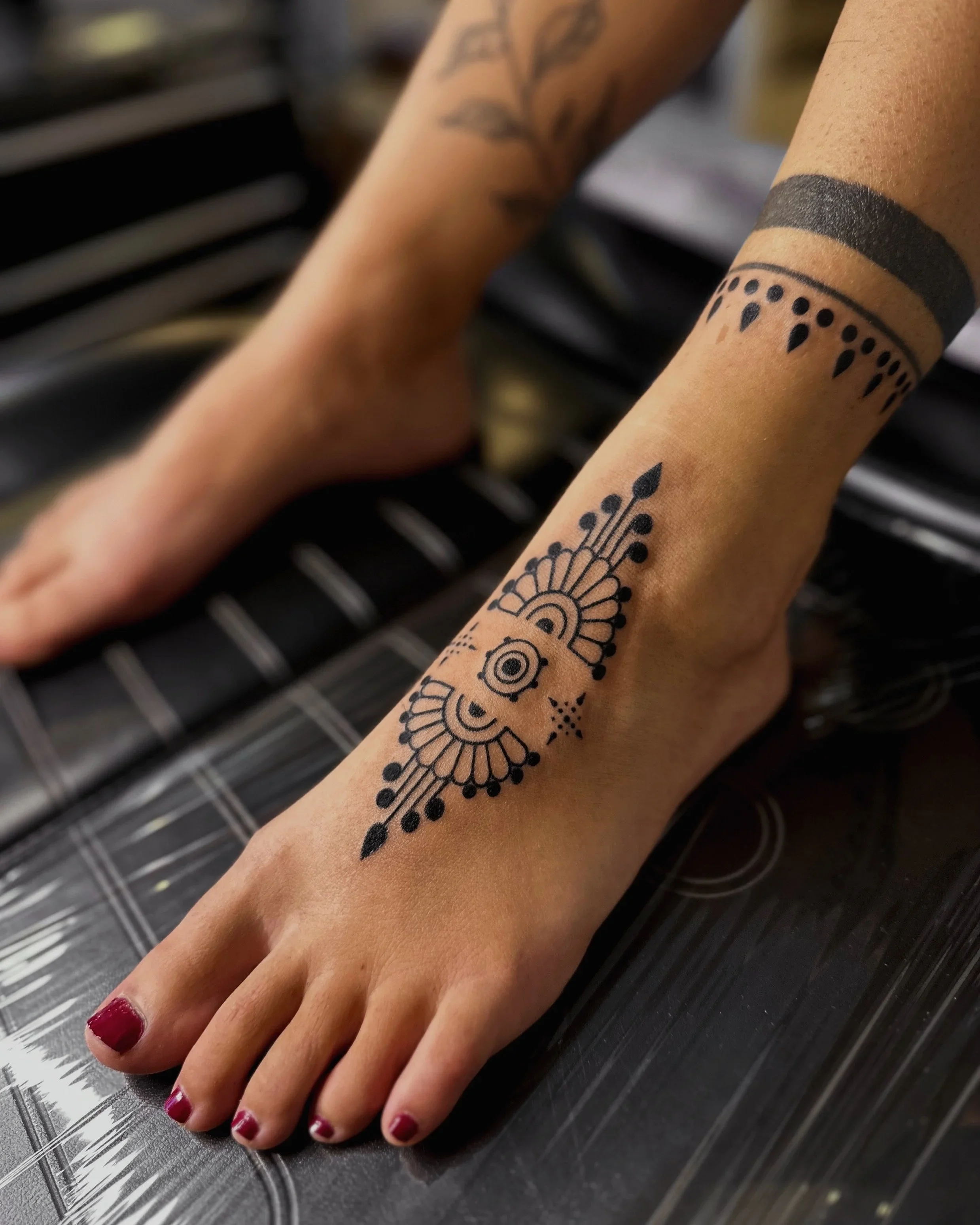 A hand with painted fingernails resting on a black, textured surface. The wrist features a black, wide band tattoo and a detailed black tattoo design resembling a mandala and floral pattern extending onto the forearm.