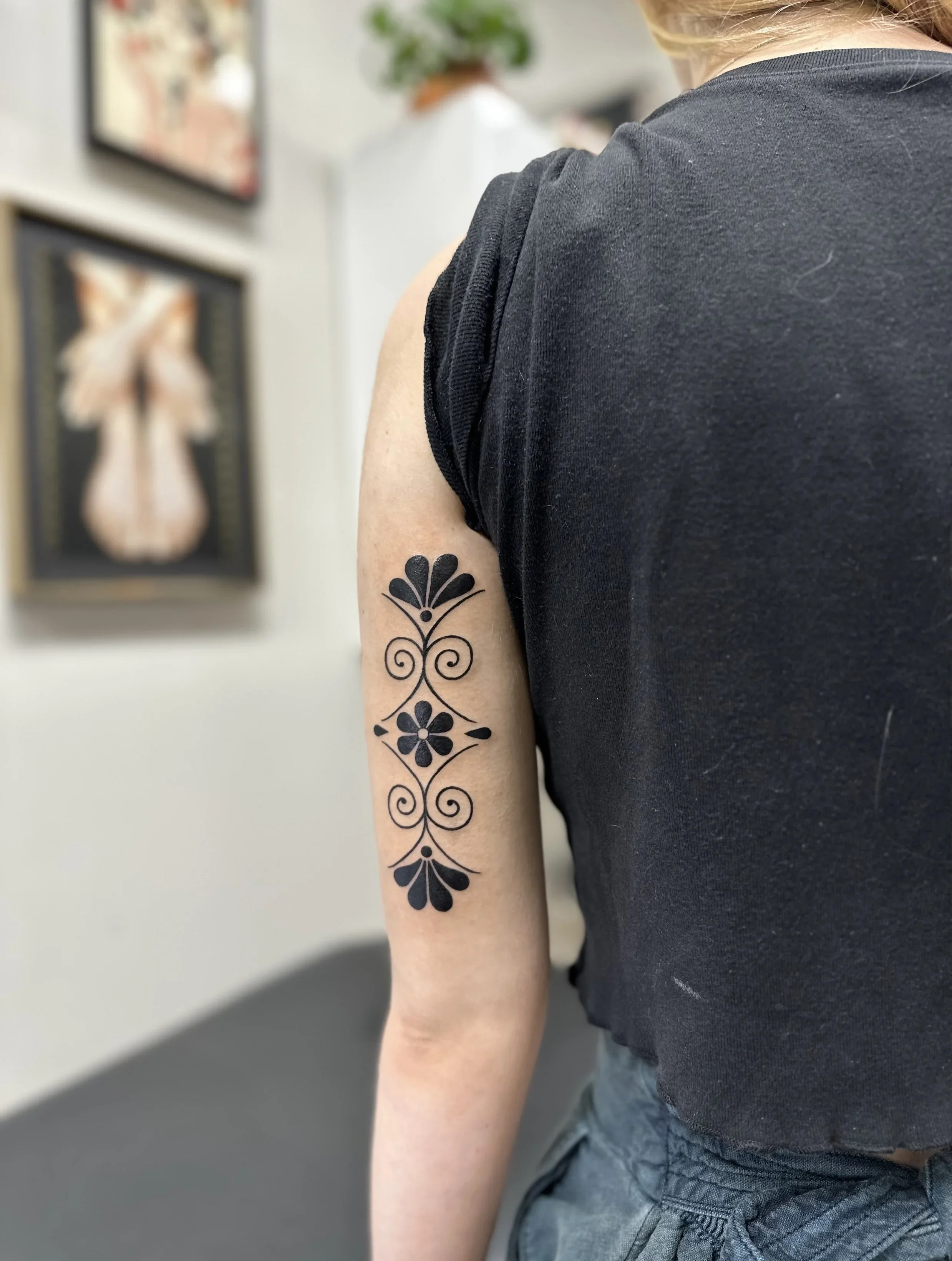 A person wearing a black sleeveless shirt and gray shorts, with a floral tattoo on their upper arm, is shown from the side. In the background, there are framed pictures and a potted plant on a shelf.