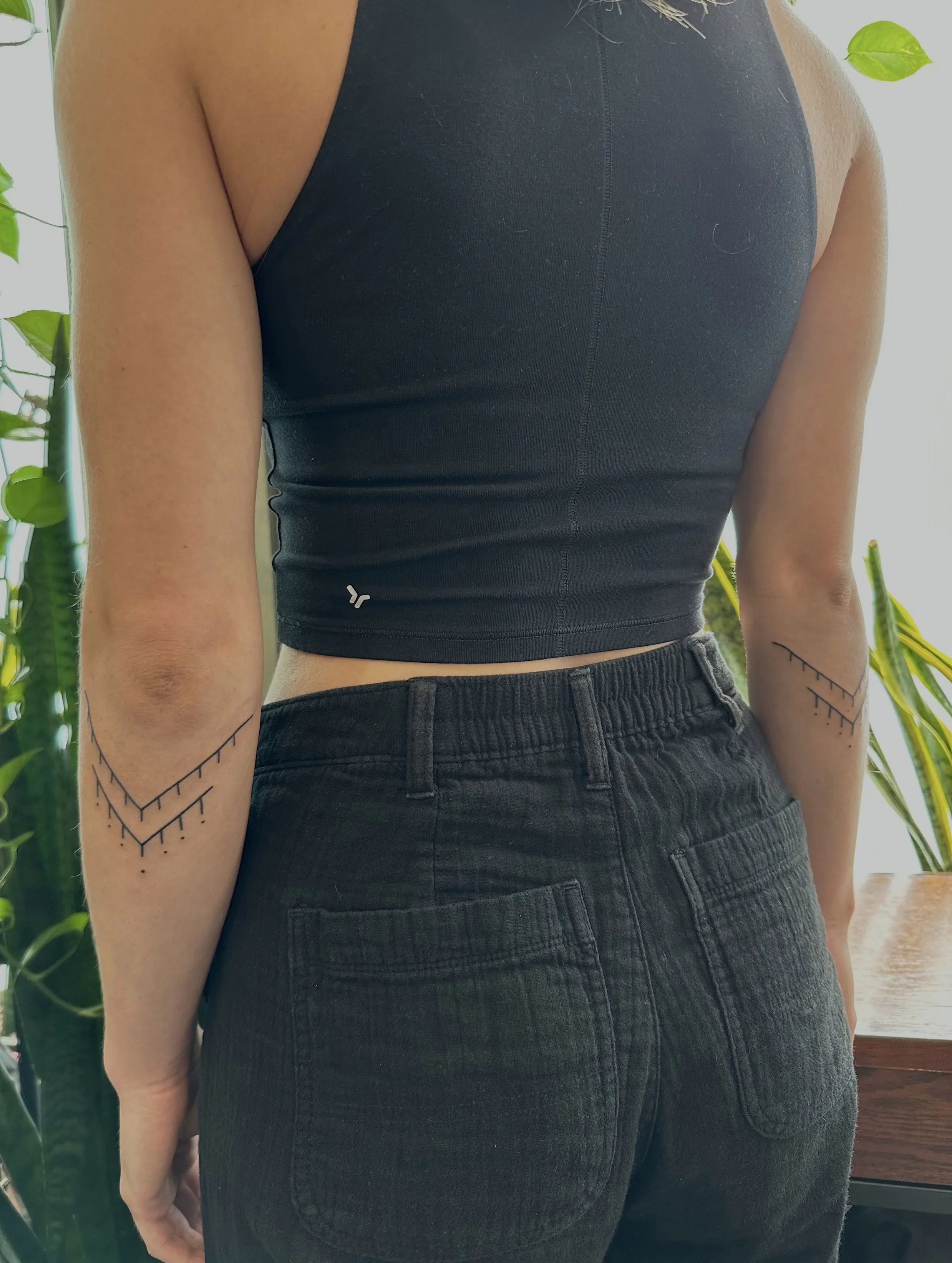 A person wearing a black sleeveless top and black jeans, with their arm tattooed with a geometric pattern. The background includes green plants and a window.