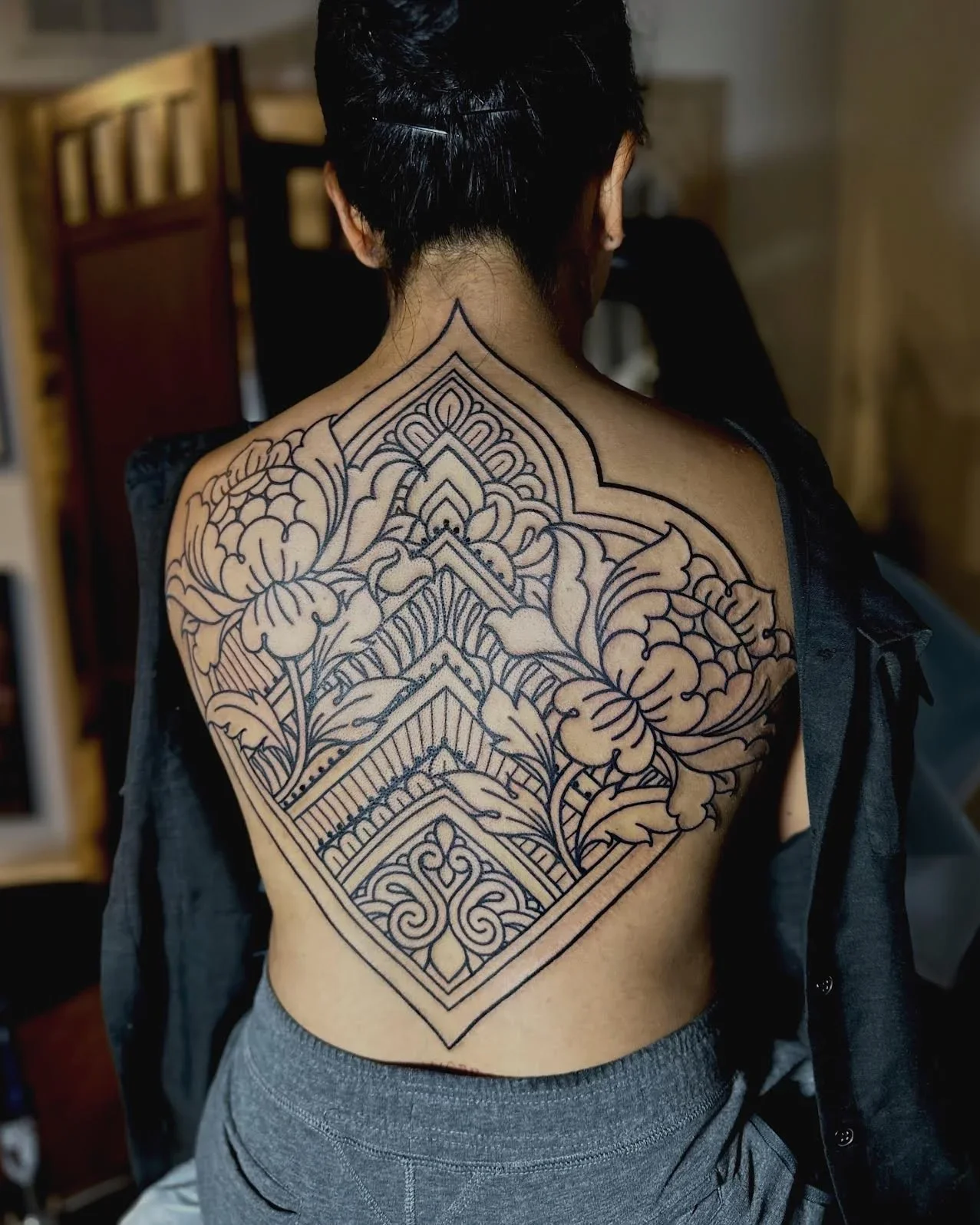 Person with a large, intricate black outline tattoo covering their back, featuring floral and geometric patterns.