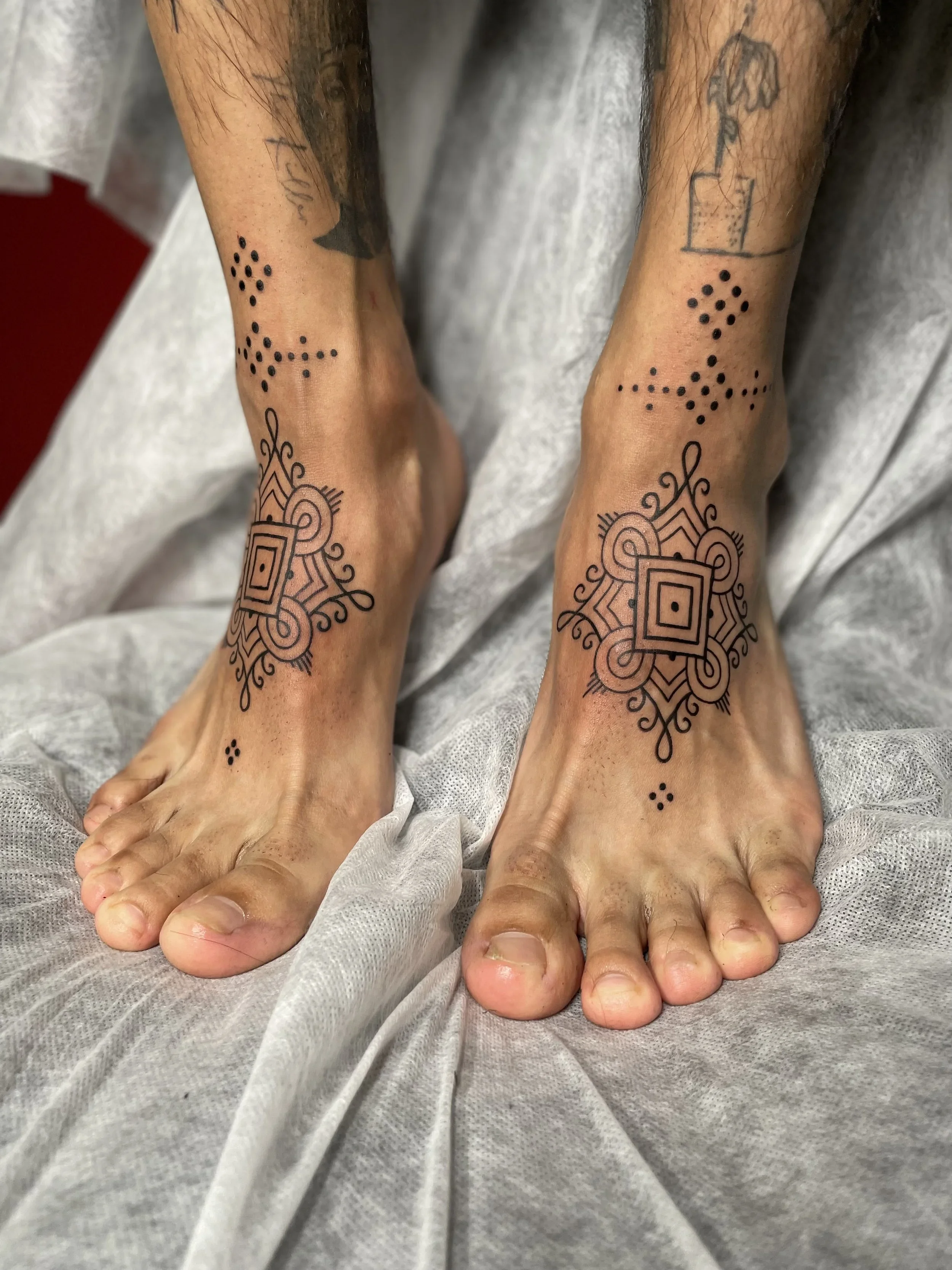 Close-up of a person's feet and ankles with intricate black tattoo designs on the skin, resting on a white fabric surface.