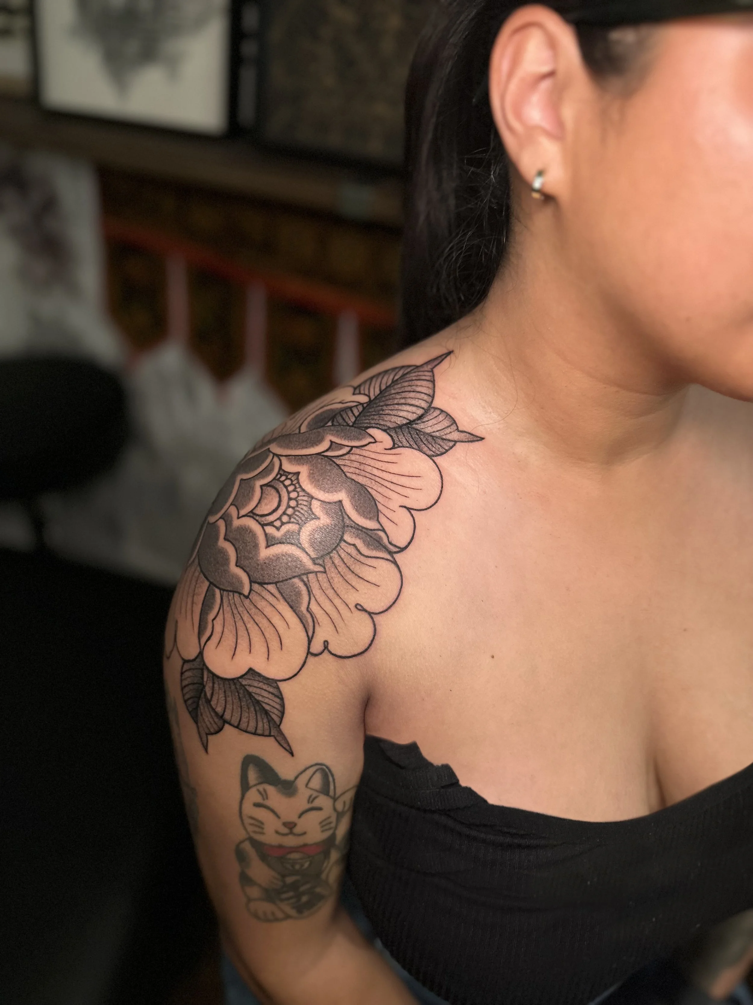 A woman with short dark hair and earrings, wearing a black tank top, displays a tattoo of a large flower on her shoulder and a cartoon cat tattoo on her elbow.