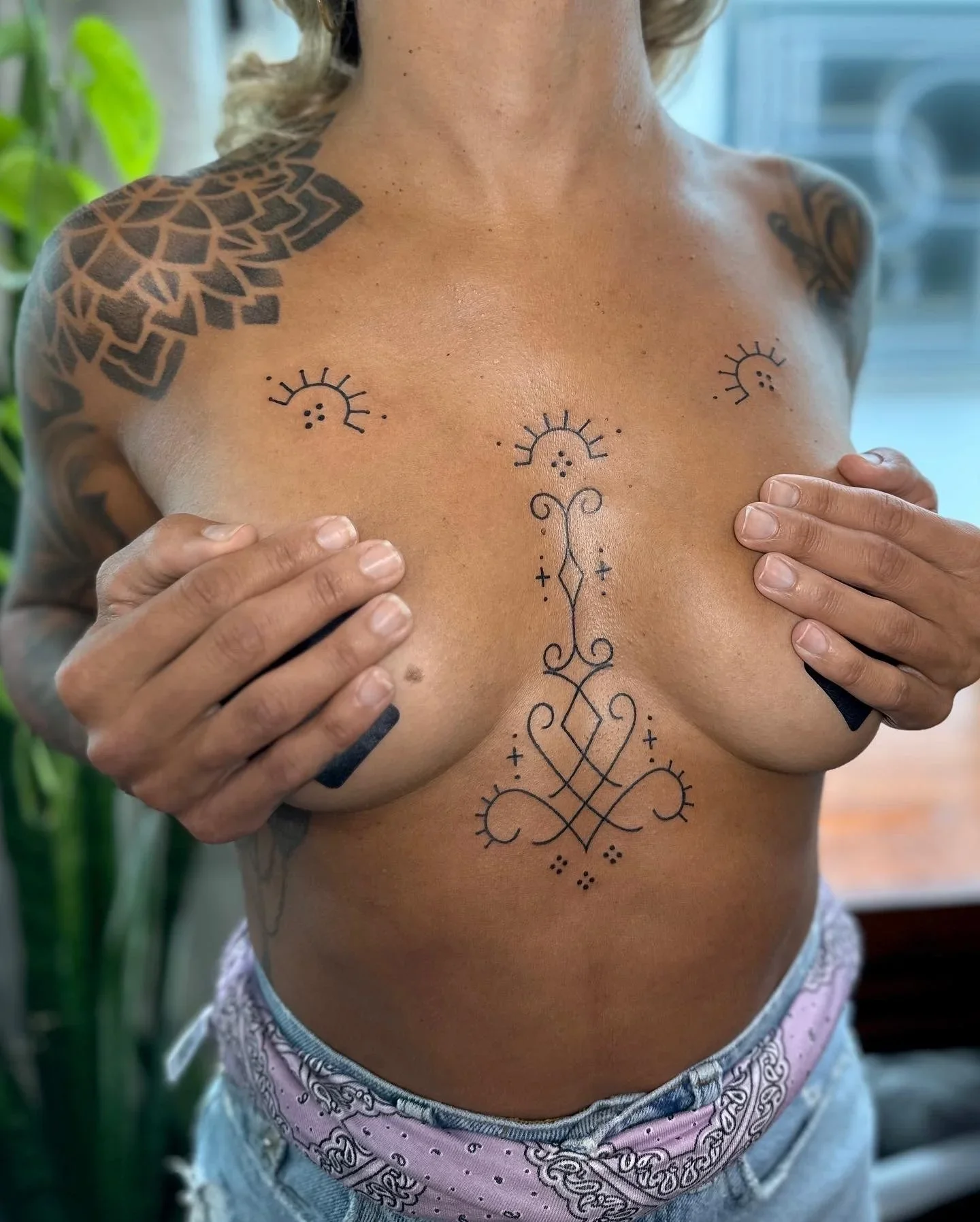 A person with tattoos on their chest and shoulders, covering their breasts with hands, in front of a plant and a window, wearing patterned shorts.