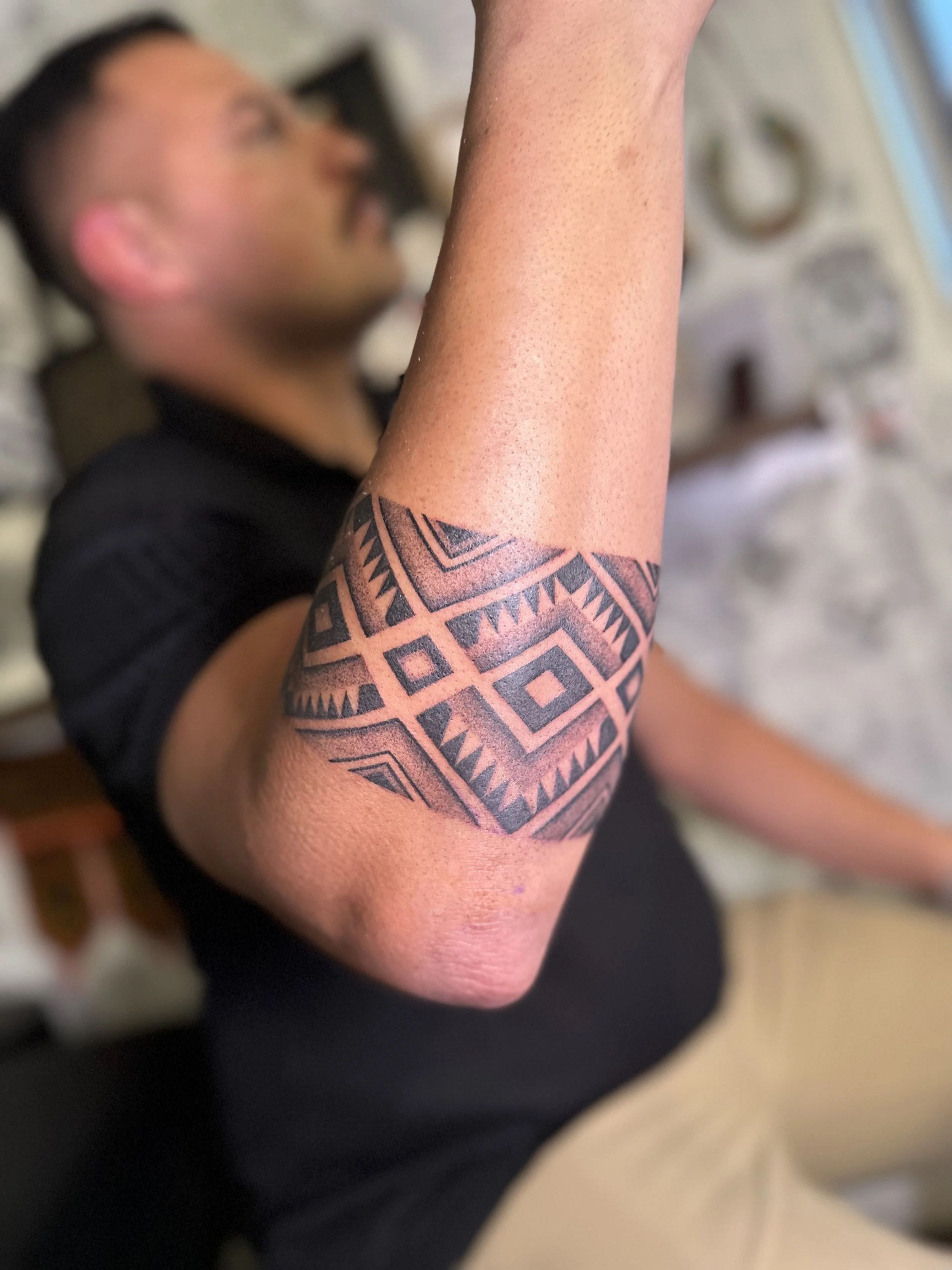 Close-up of an arm with a detailed geometric black tattoo, with a person lying down in the background, out of focus.