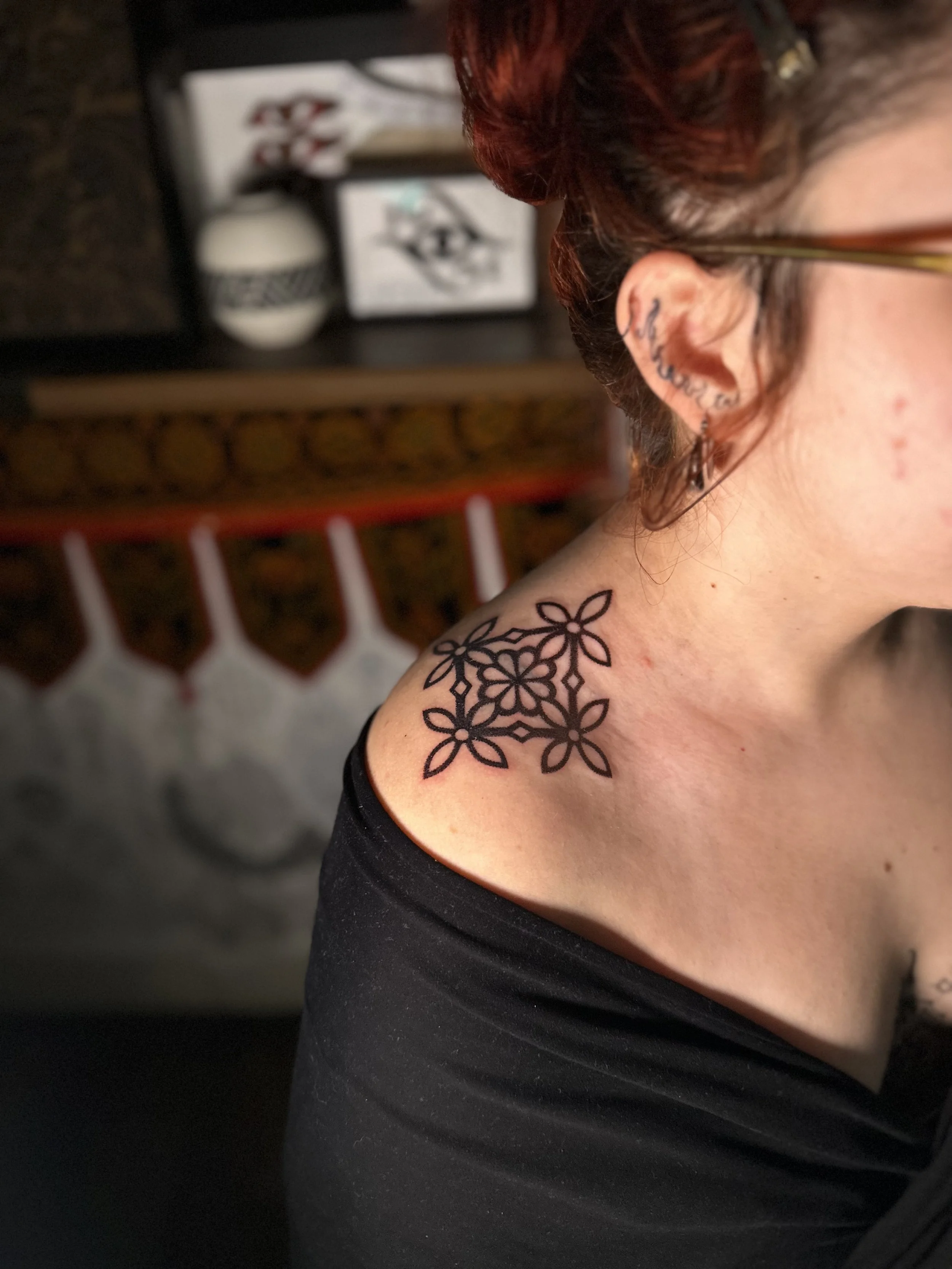 Close-up of a person's shoulder and neck with a floral tattoo, wearing a black off-the-shoulder top, and earrings. A blurred background includes a white vase and framed artwork.