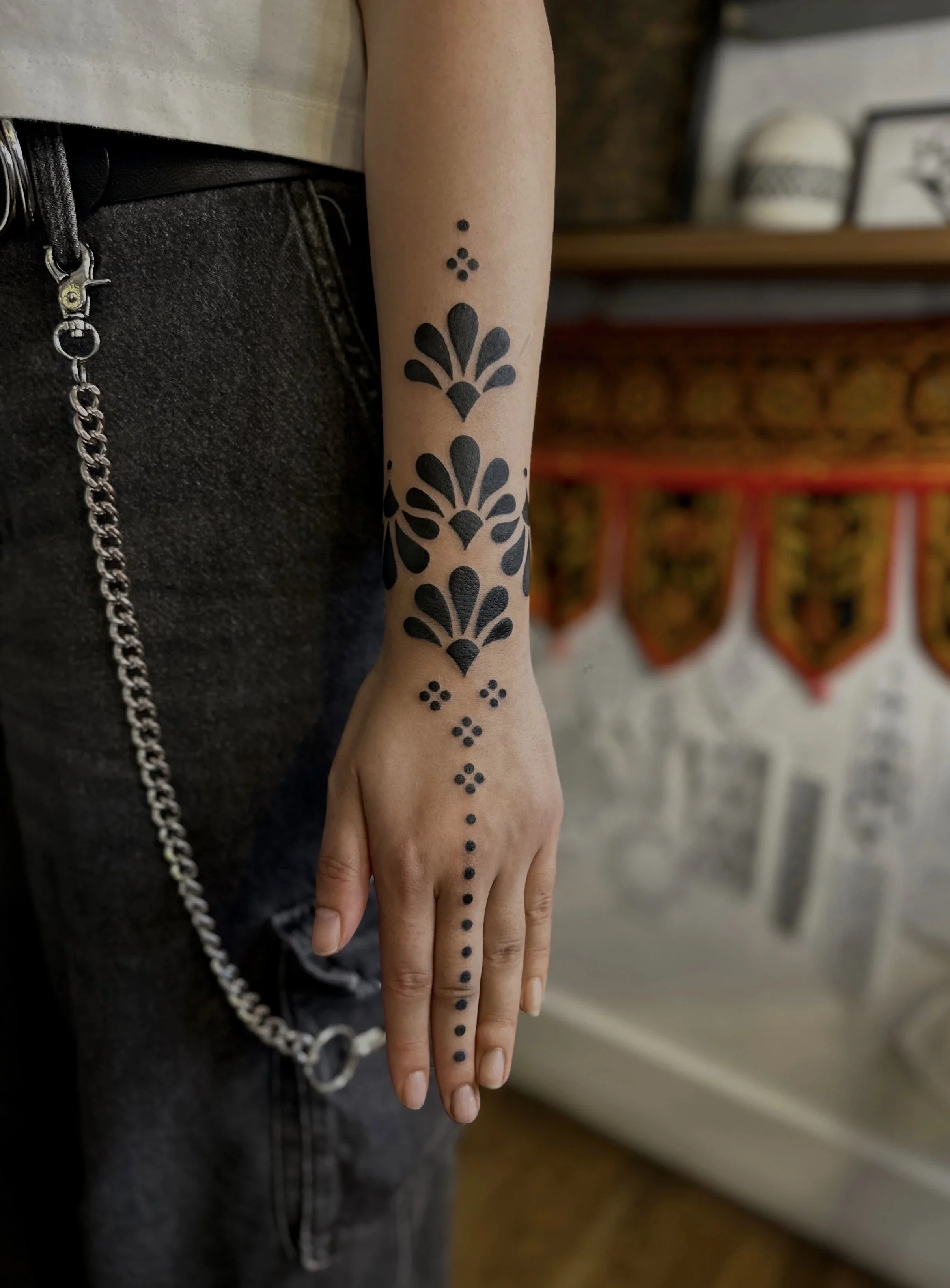 A person's arm and hand with black floral-style temporary tattoos in a vertical pattern down the forearm and hand, with small dotted accents, standing inside a shop or studio with blurred background.