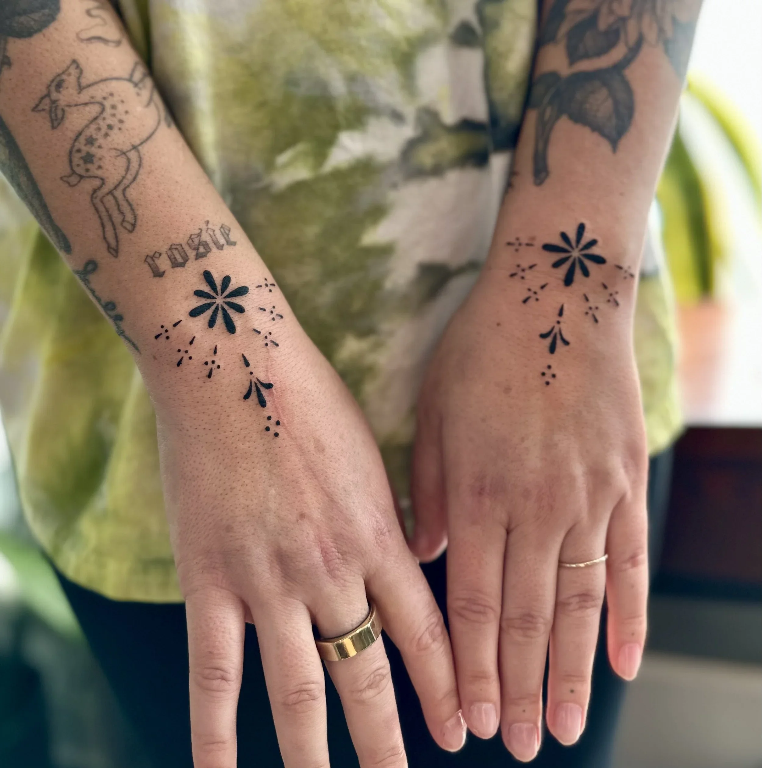Two hands with tattoos and rings resting on top of each other, with a blurred background.