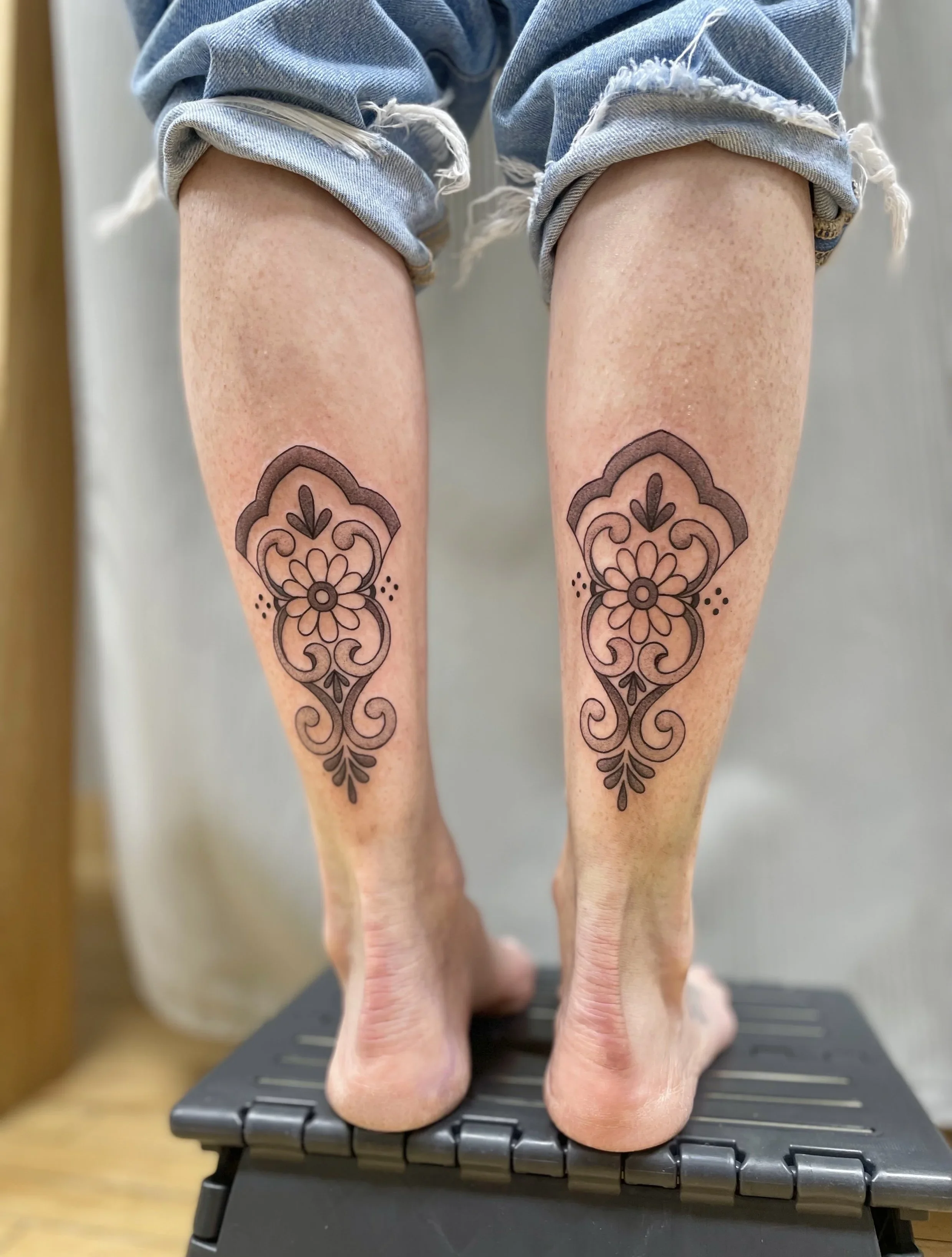 Close-up of a person's legs and feet with tattoos on the calves, standing on a black backplate with hinges, possibly on a weighing scale or medical device.