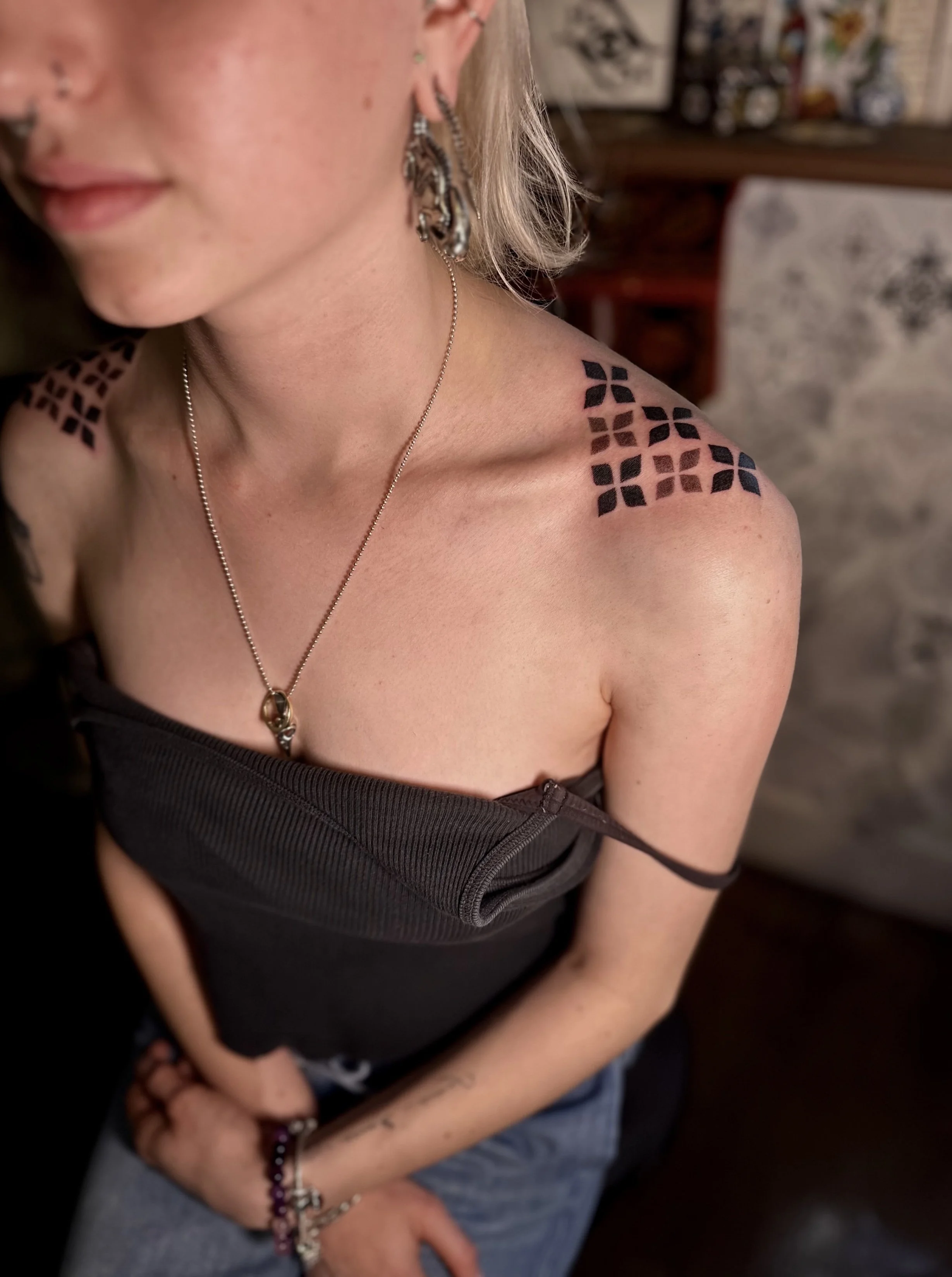 Close-up of a young woman with tattoos on her shoulder and arm, wearing earrings, a necklace, and a black off-shoulder top, standing indoors with blurry background.