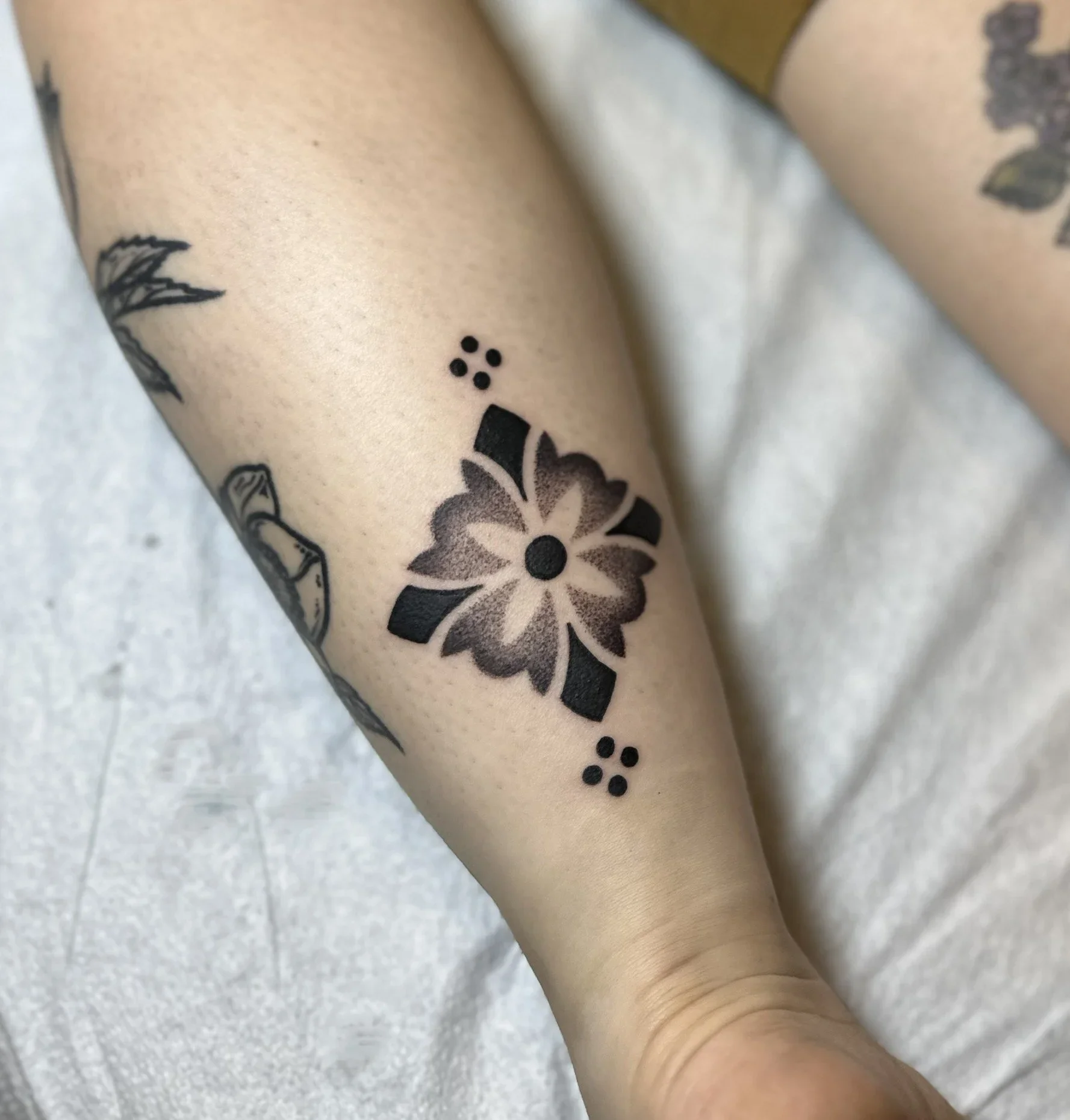 Close-up of a person's arm with several black tattoos, including a flower, dots, and geometric shapes.