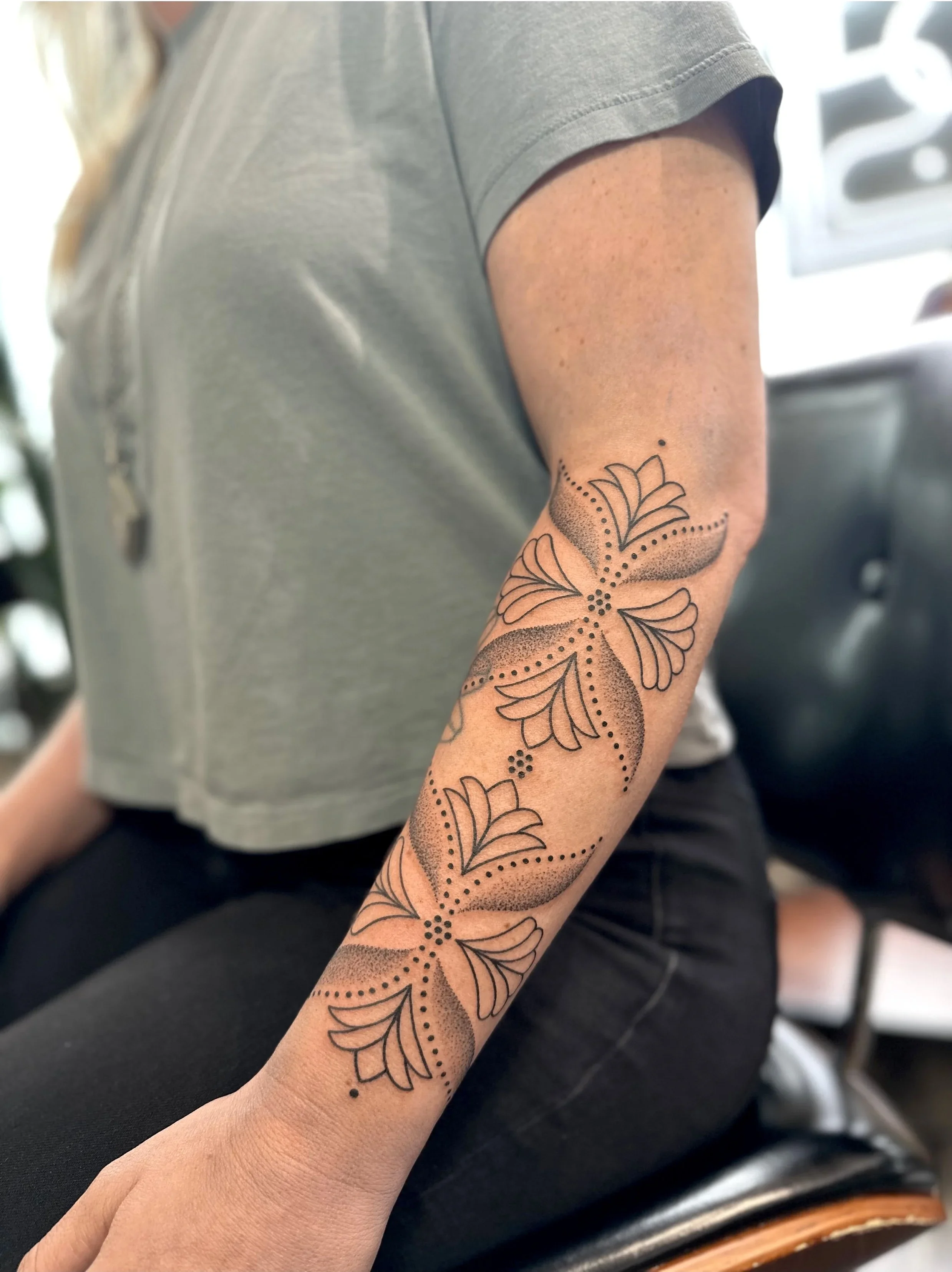 A person's arm with a floral tattoo featuring leaves and dots on their forearm, wearing a gray short-sleeve shirt.