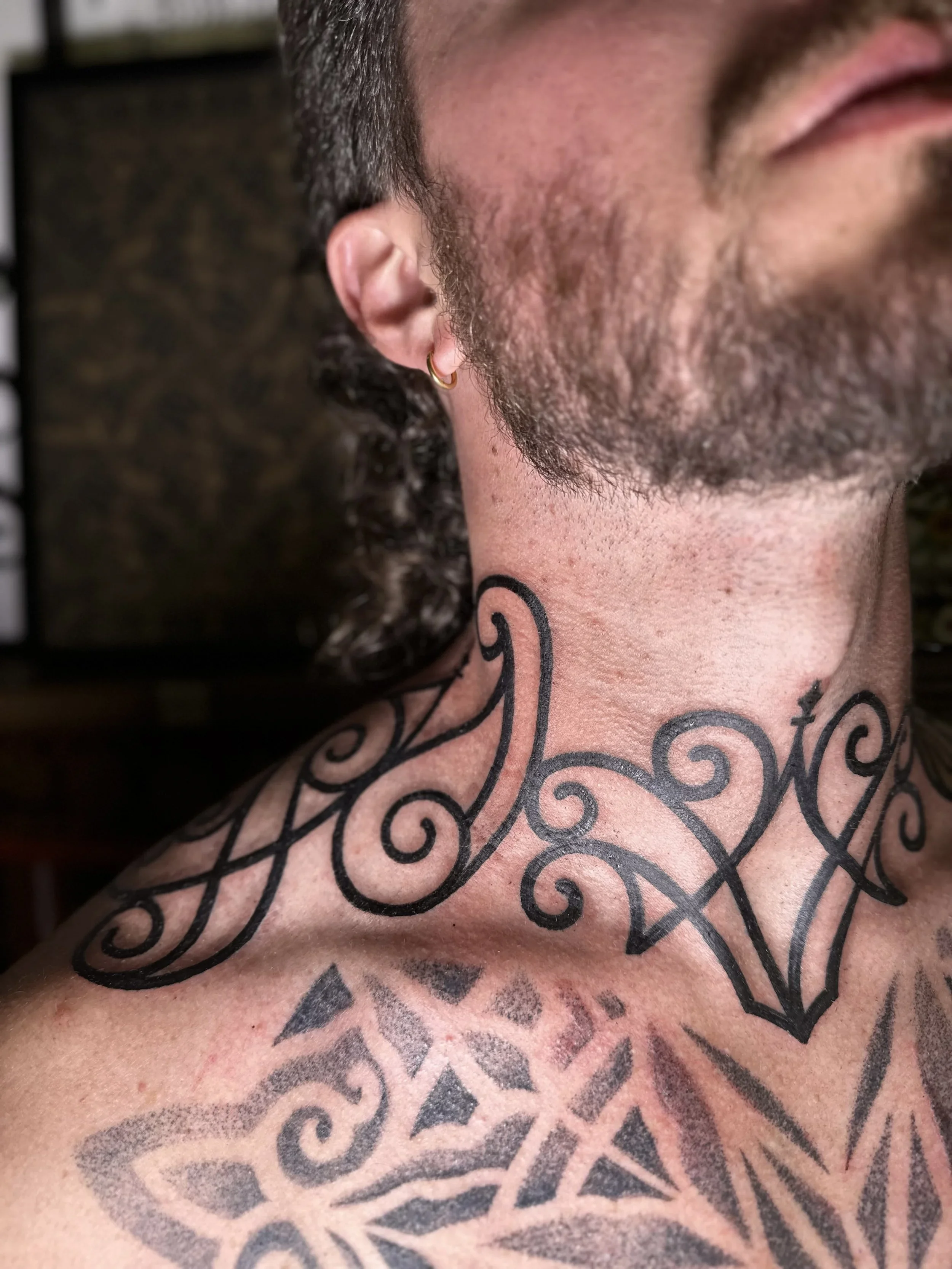 Close-up of a person's cheek, ear, and beard, with a large black tattoo of intricate swirling patterns and shapes covering the skin.