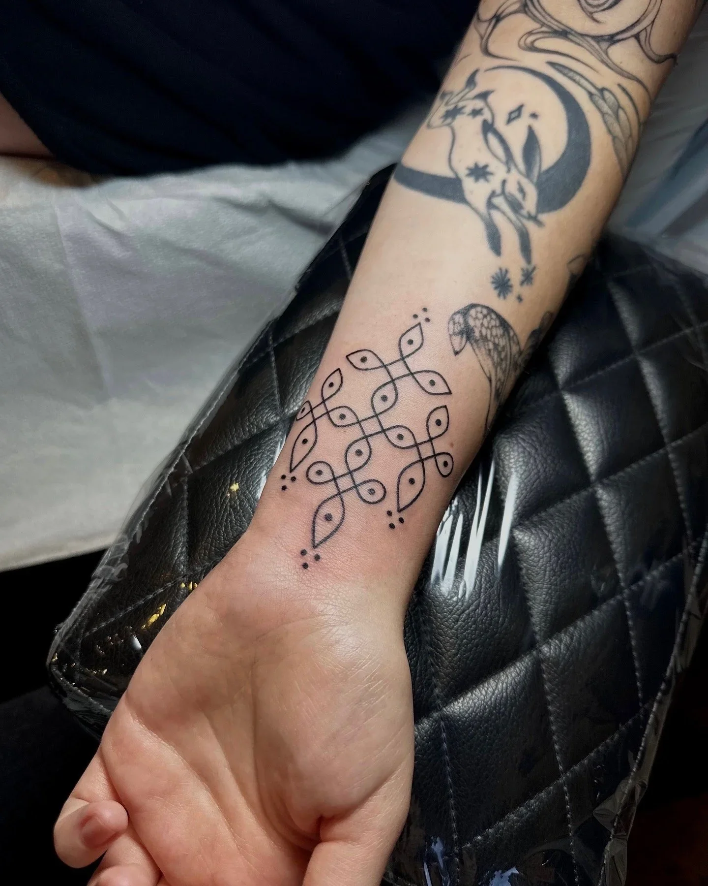 Person's arm with several tattoos, including a crescent moon with stars, a geometric pattern, and a snake, resting on a black quilted surface.