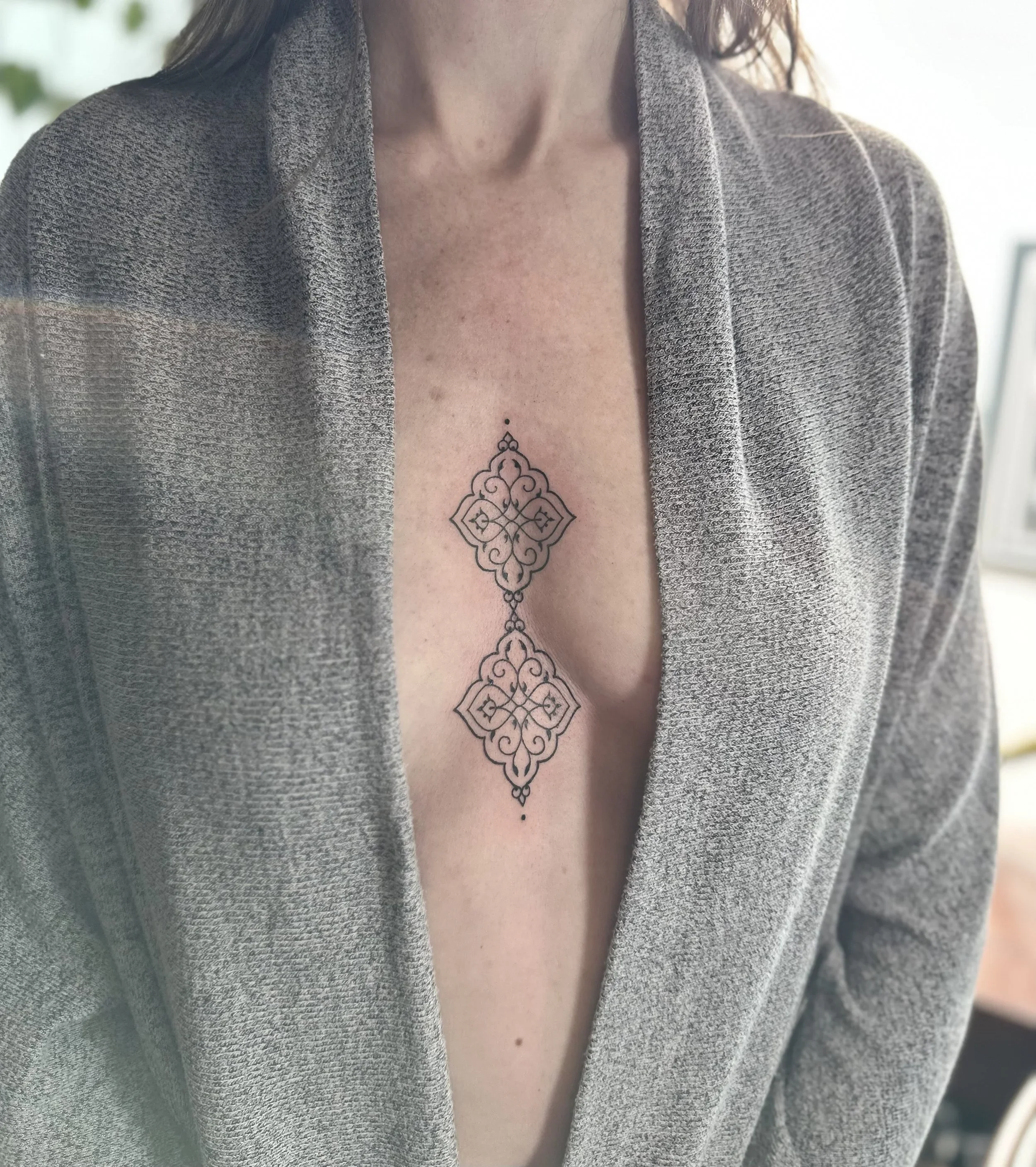 Close-up of a woman wearing a gray textured blazer, showing her chest with a tattoo of an ornate symmetrical geometric design.