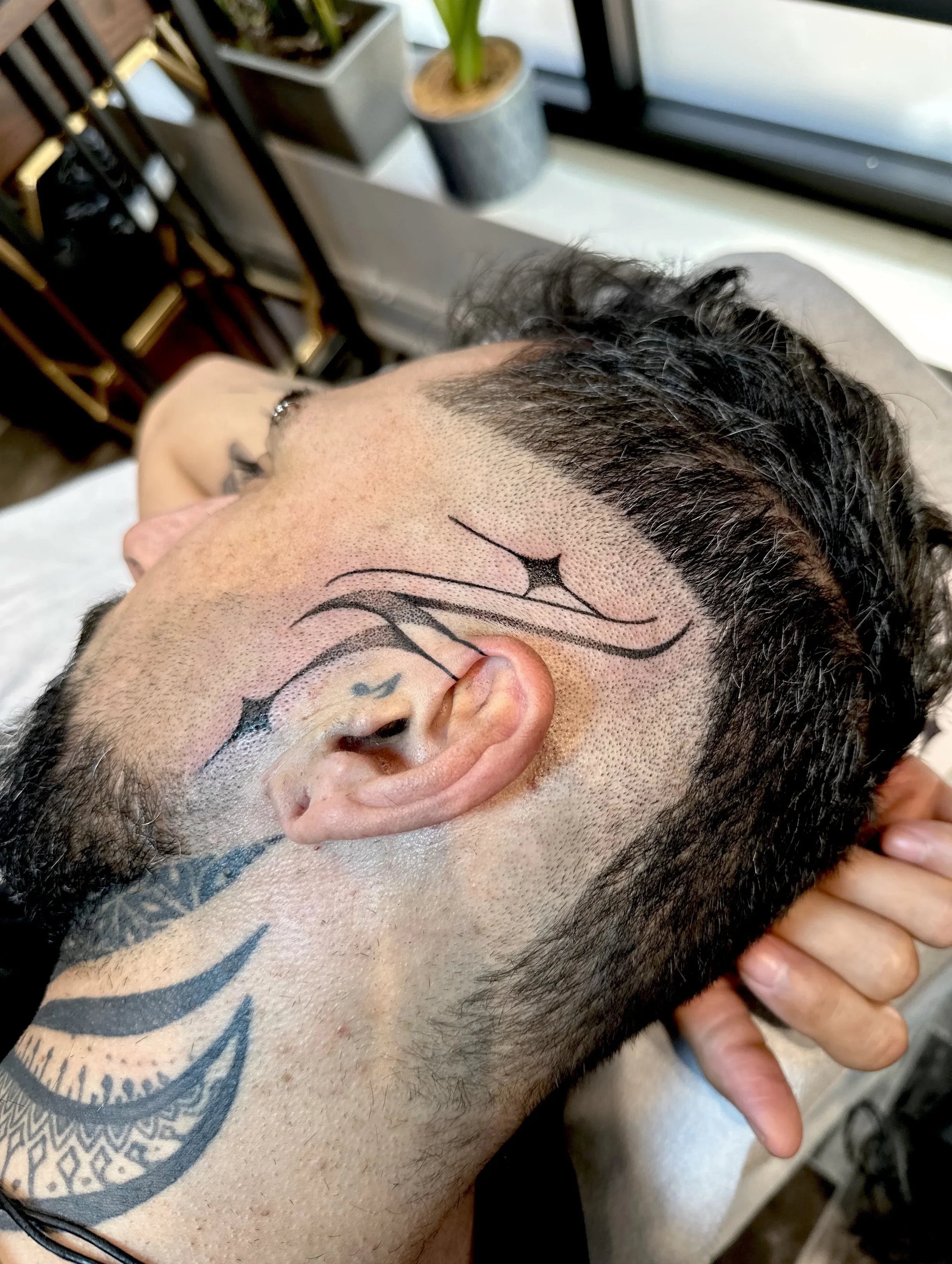 A person with tattoos on the neck and chest, lying on their side with head resting on a surface near a window, showing fresh facial tattoo lines.