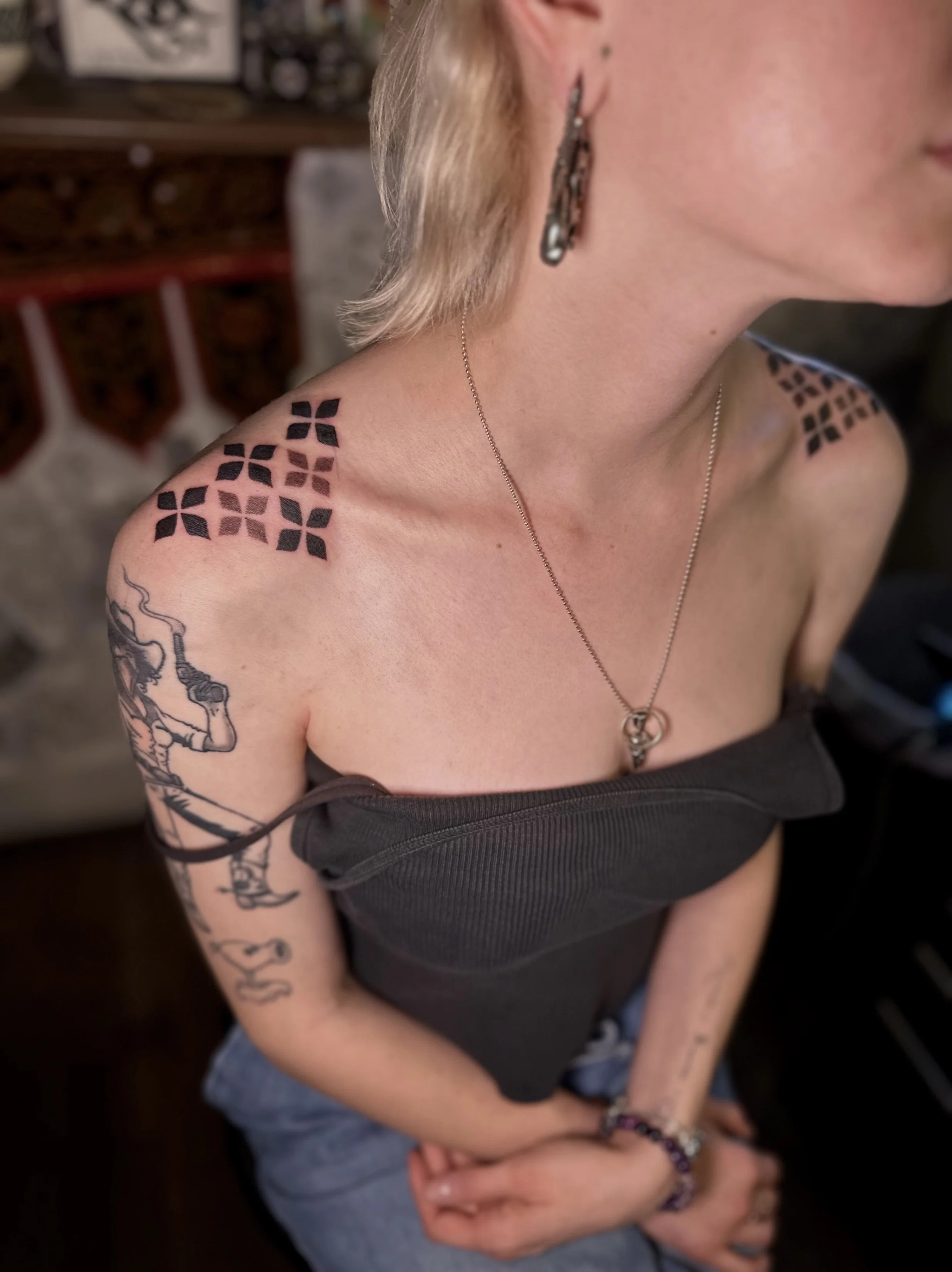 A woman with tattoos on her shoulder and arm, wearing earrings and a necklace, dressed in a black strapless top, sitting with crossed arms in a room with a dark background.