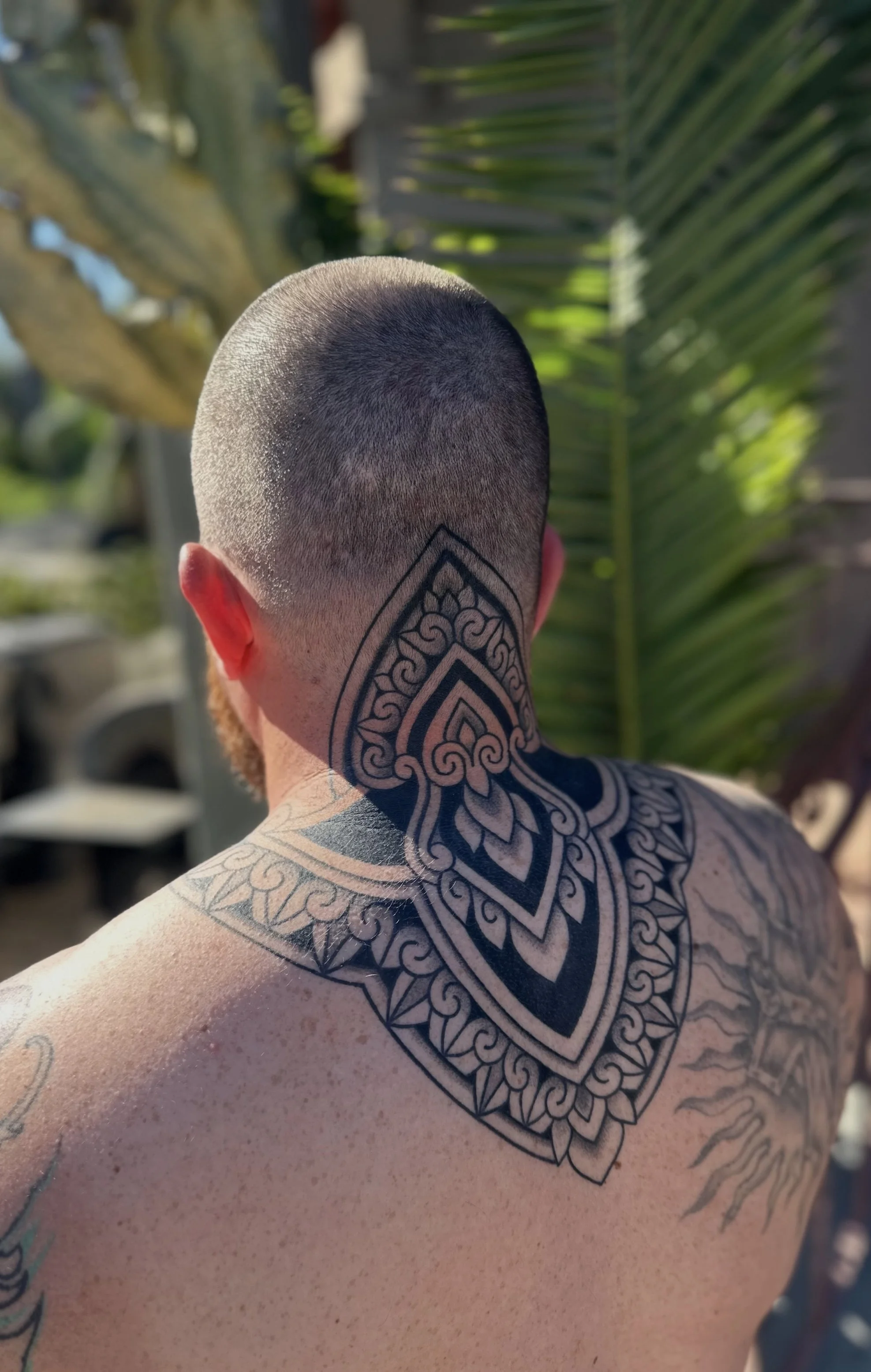 Back of a person's head with a shaved hairstyle and a geometric tattoo on the neck and shoulder, outdoors with green foliage in the background.