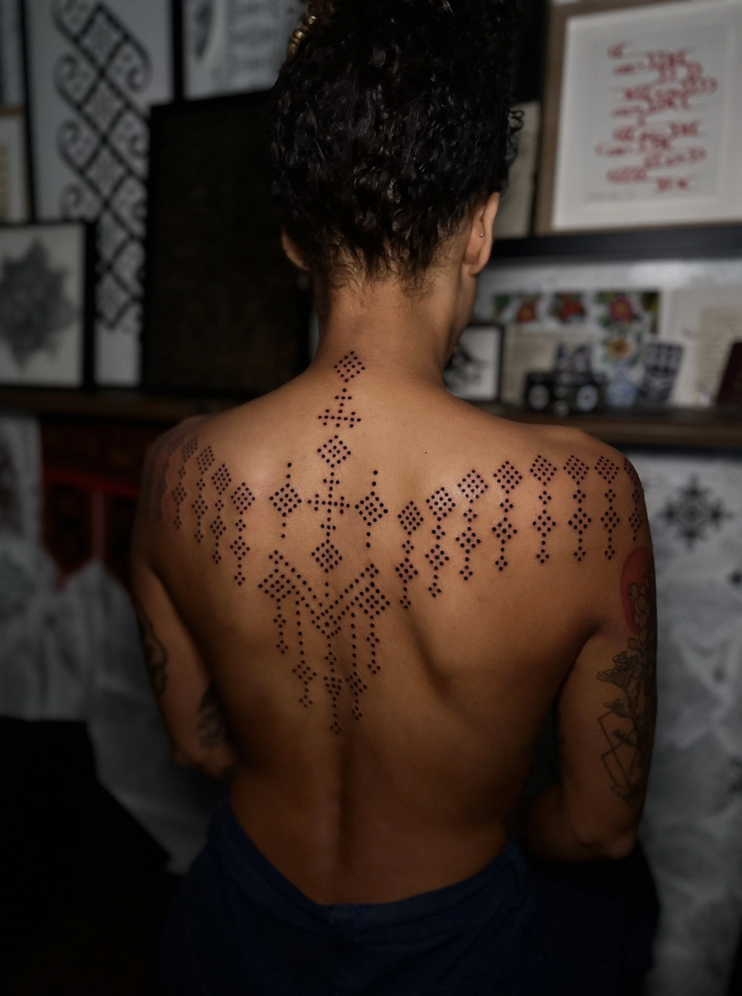 Rear view of a person with short curly hair, showing geometric black dot tattoo patterns on their neck and shoulders, in an indoor setting with artwork visible on the wall behind.