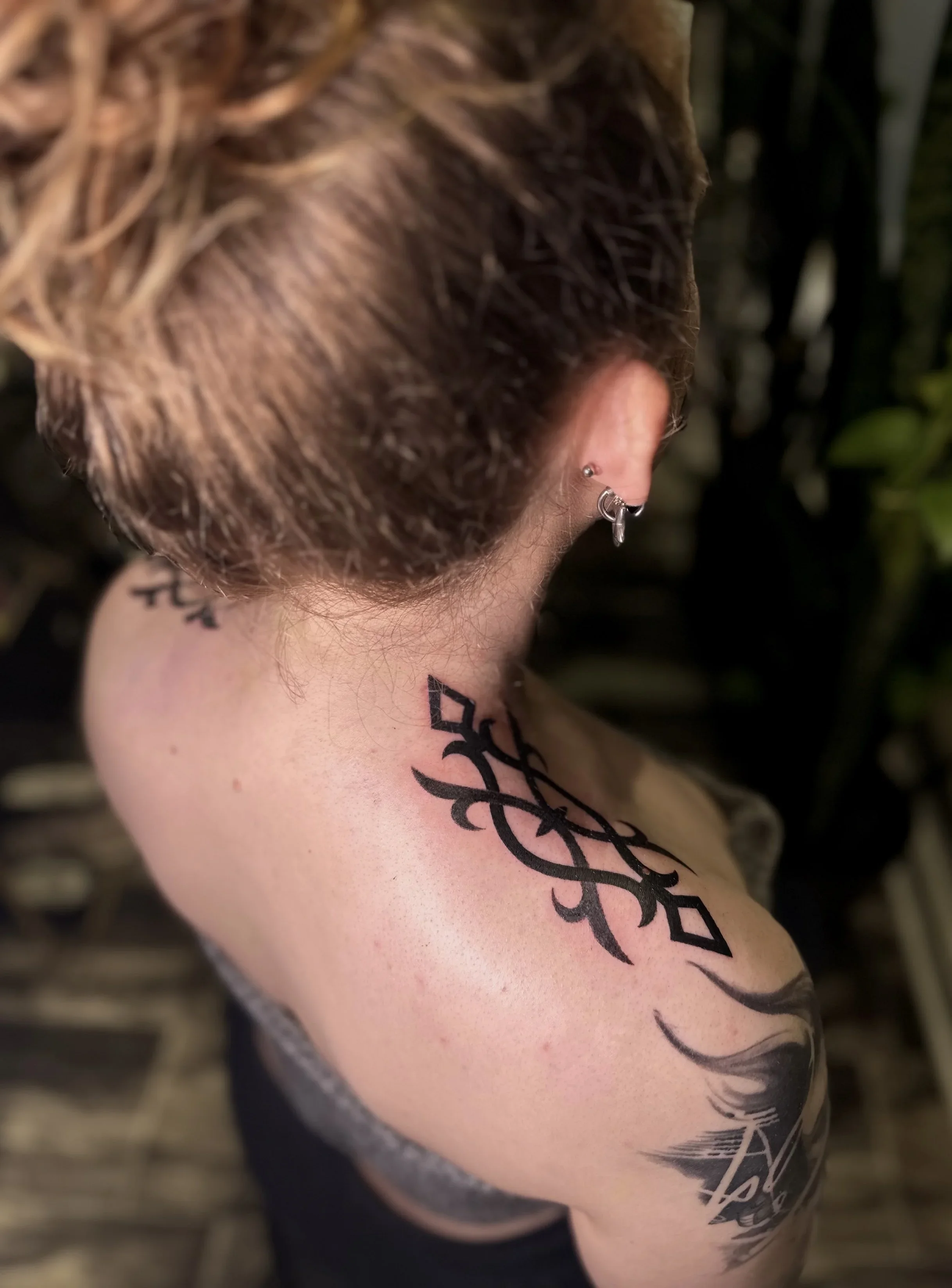 A person with short, wavy, blonde hair has multiple earrings in their ear and a tattoo on their shoulder and upper arm. The tattoo includes words and decorative designs.