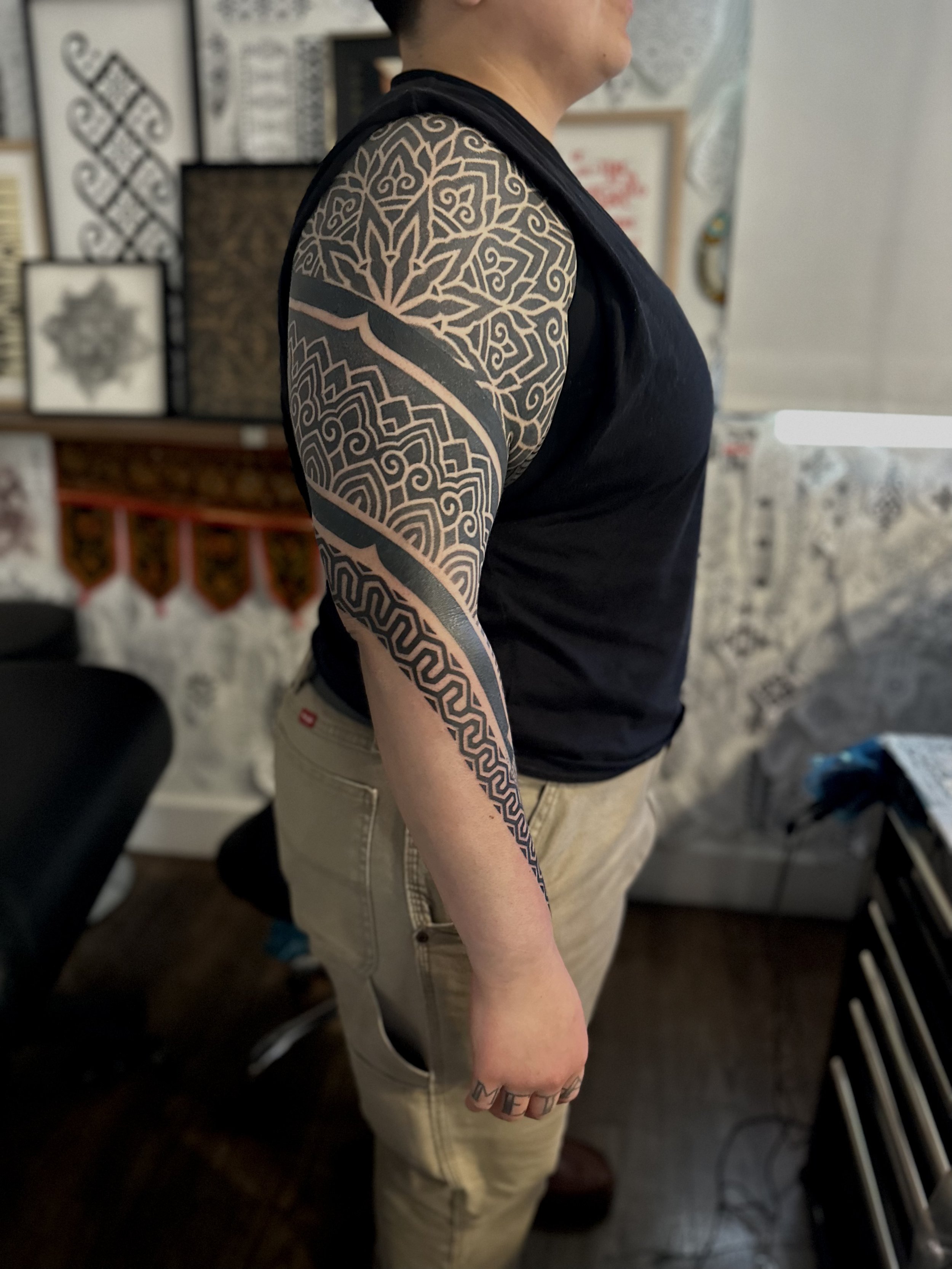 A person with a tattoo sleeve on their arm, standing indoors in front of framed artwork and decorations.