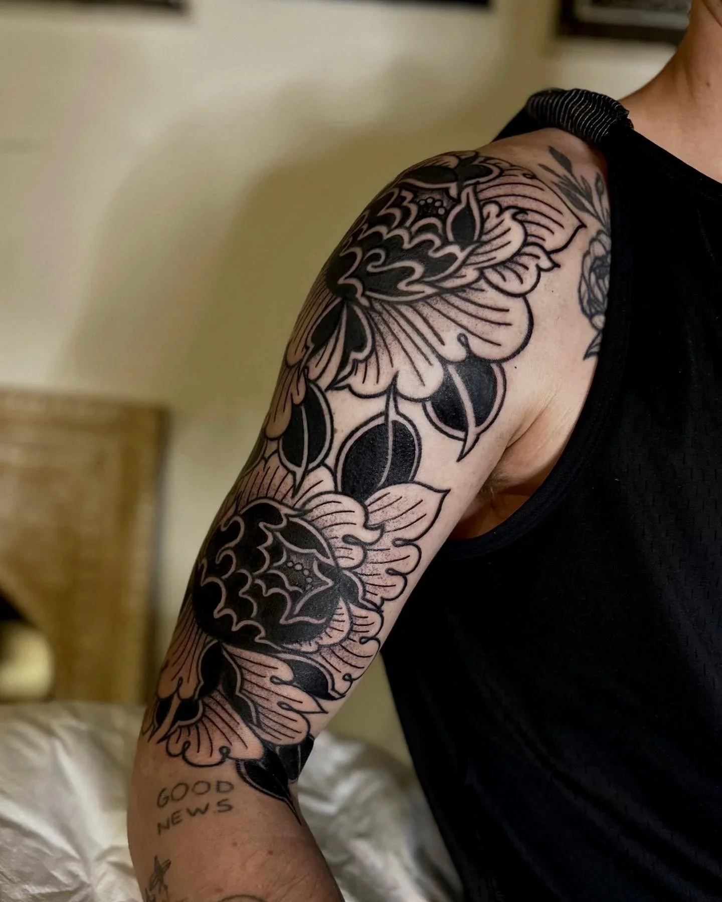 Close-up of a person's upper arm showing a black and grey floral tattoo with a sun and wave design. The person is wearing a black sleeveless shirt and a black wristband. The background is blurred, and there is a paper underneath their arm. Near the w