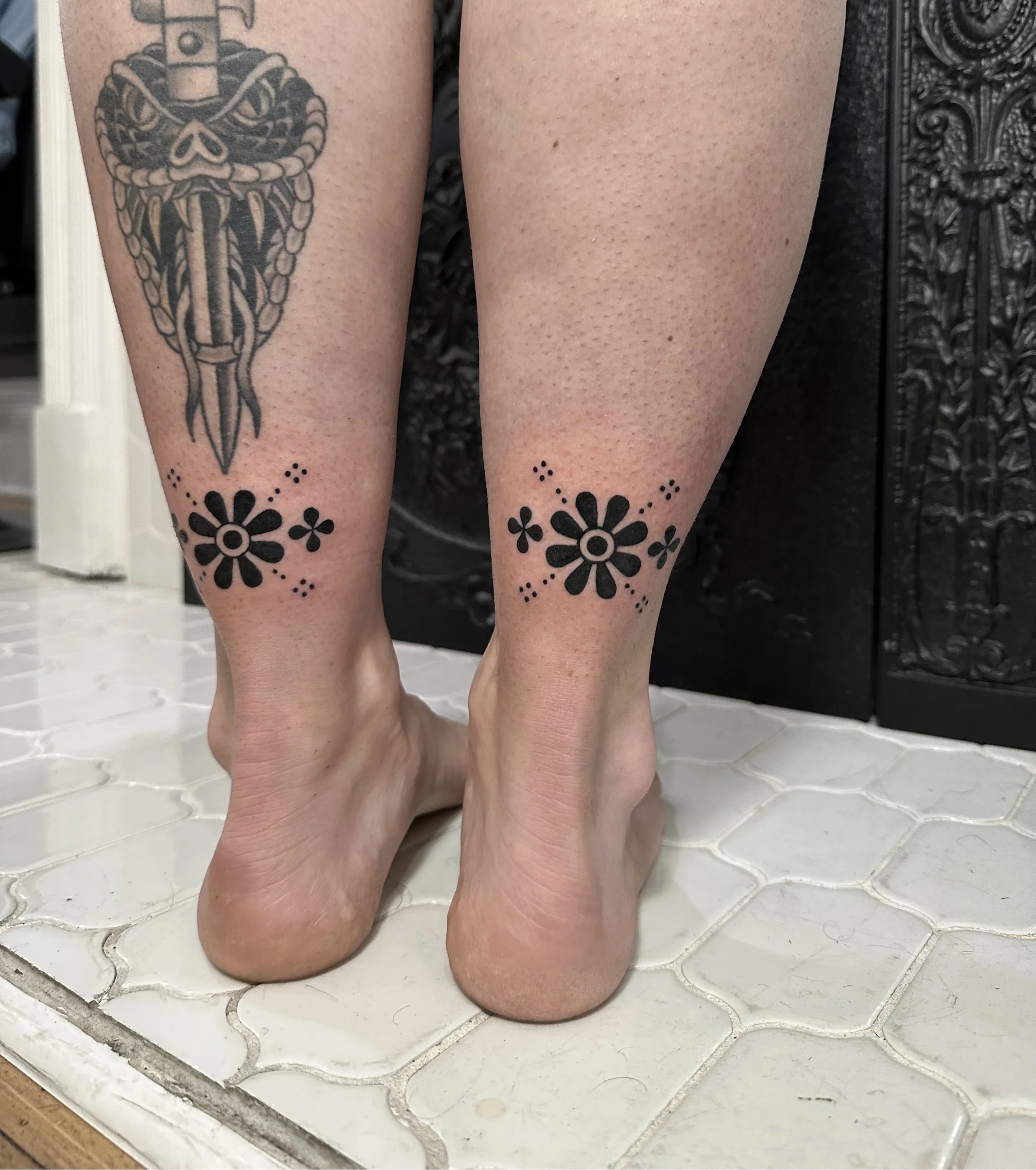 Close-up of a person's lower legs and feet with floral tattoos on the calves, standing on a white tiled surface with a cracked pattern. The background includes a decorative black metal object.