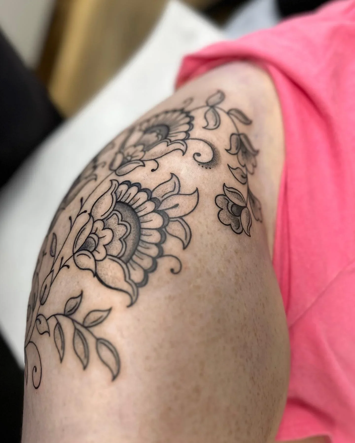 Close-up of a person's arm with an intricate black and gray floral tattoo, wearing a pink top.