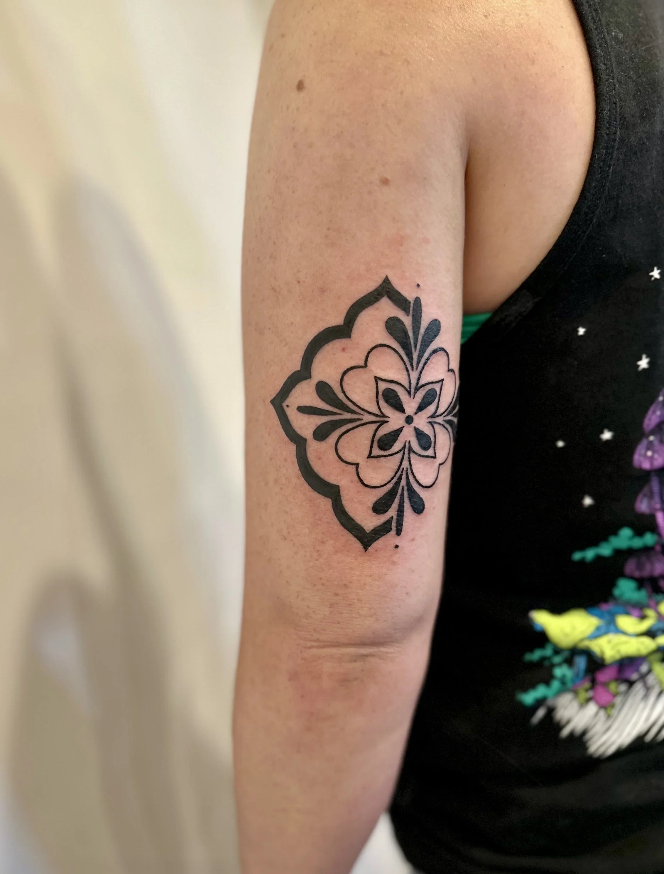 Close-up of a person's upper arm and shoulder, showing a black tattoo of a floral pattern on the inner arm.