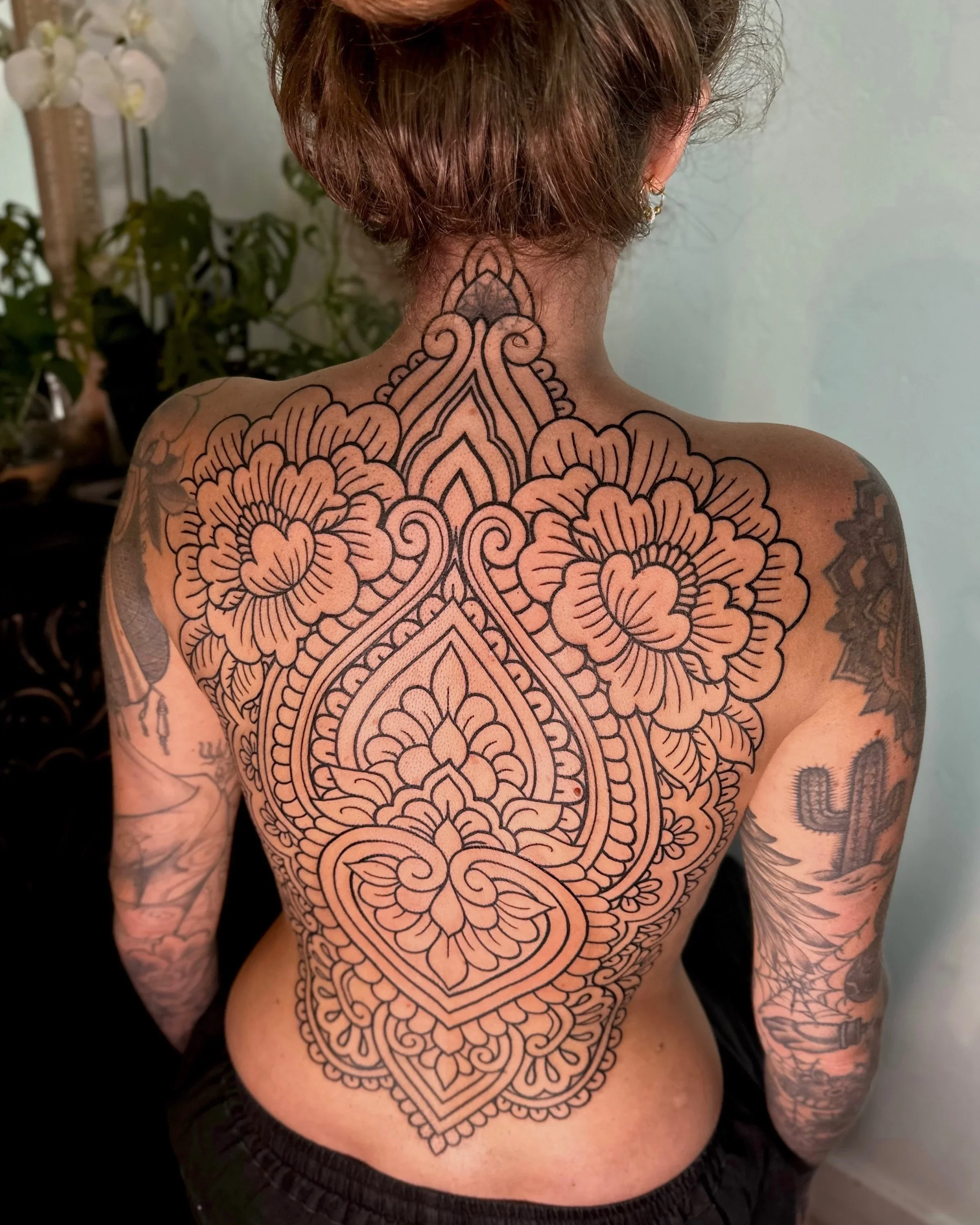 A person with shoulder-length brown hair and tattoos on their arms and back, showing a large intricate black outline tattoo on their back and neck. The tattoo features floral and geometric patterns that extend from the neck down to the lower back.