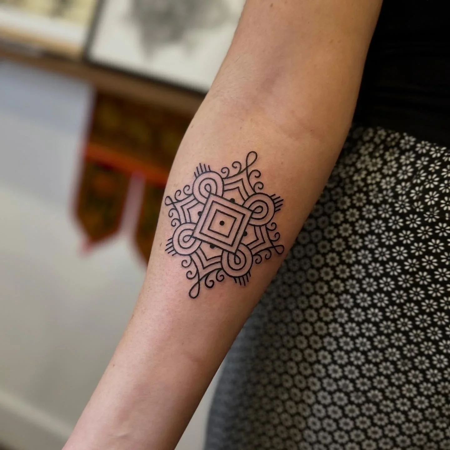 Line art geometric tattoo on person's forearm, featuring a symmetrical pattern with diamonds, circles, and swirling lines.