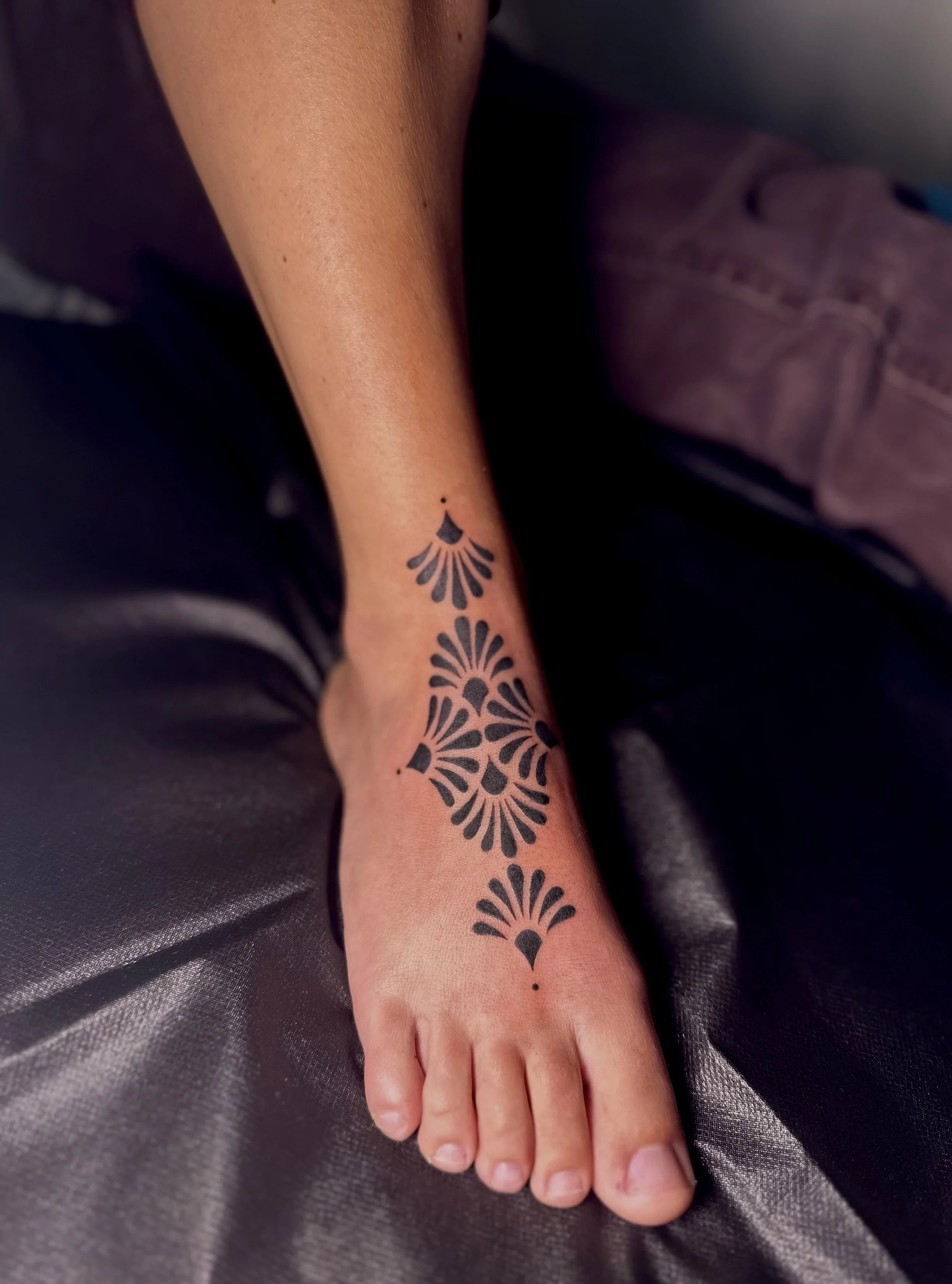 Close-up of a person's forearm and hand with a black, floral-style tattoo on the lower forearm. The person is resting their hand on a shiny, dark gray fabric surface.