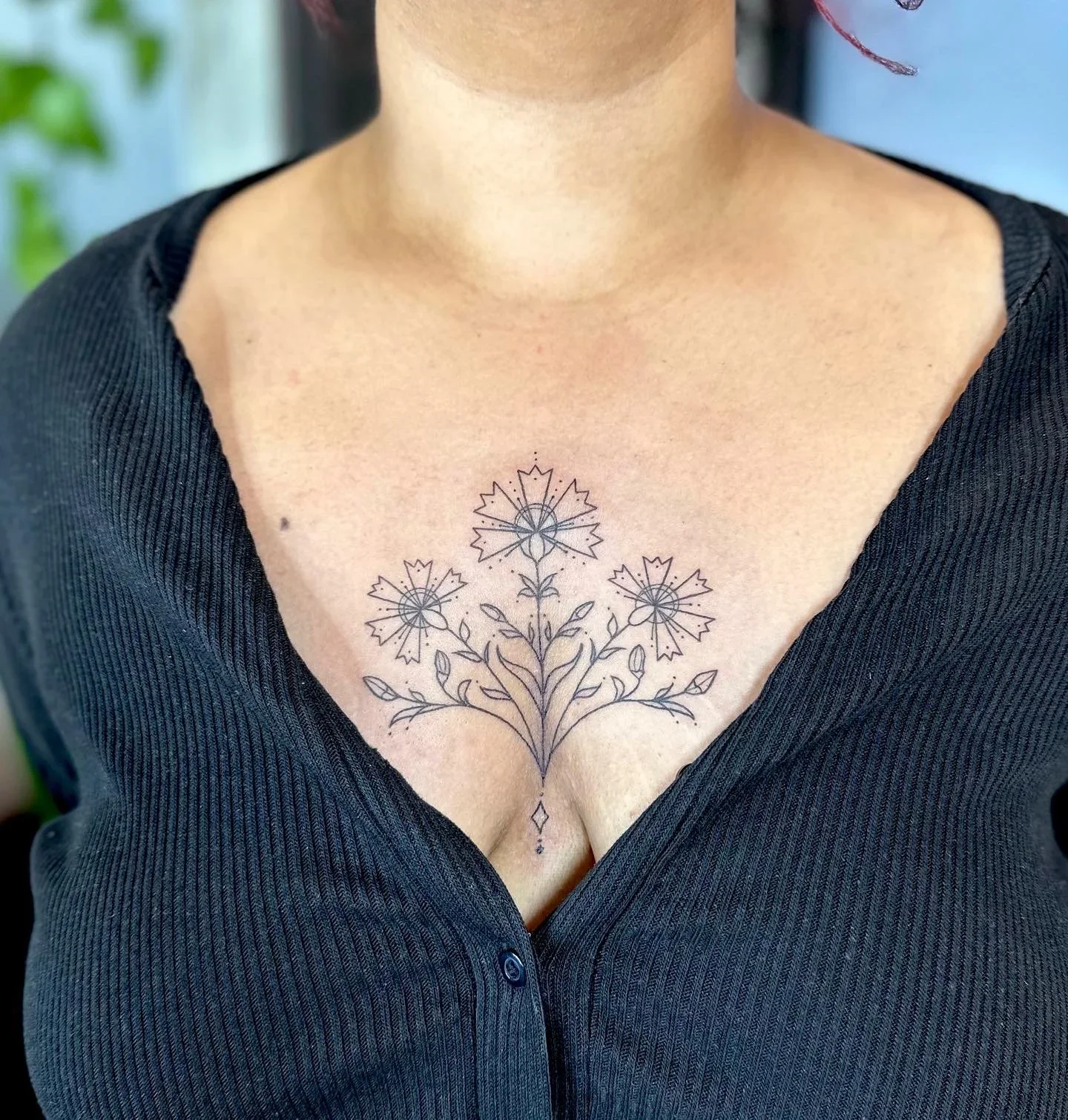 A woman with a black dress showing a floral tattoo on her chest, featuring three flowers with leaves and stems.