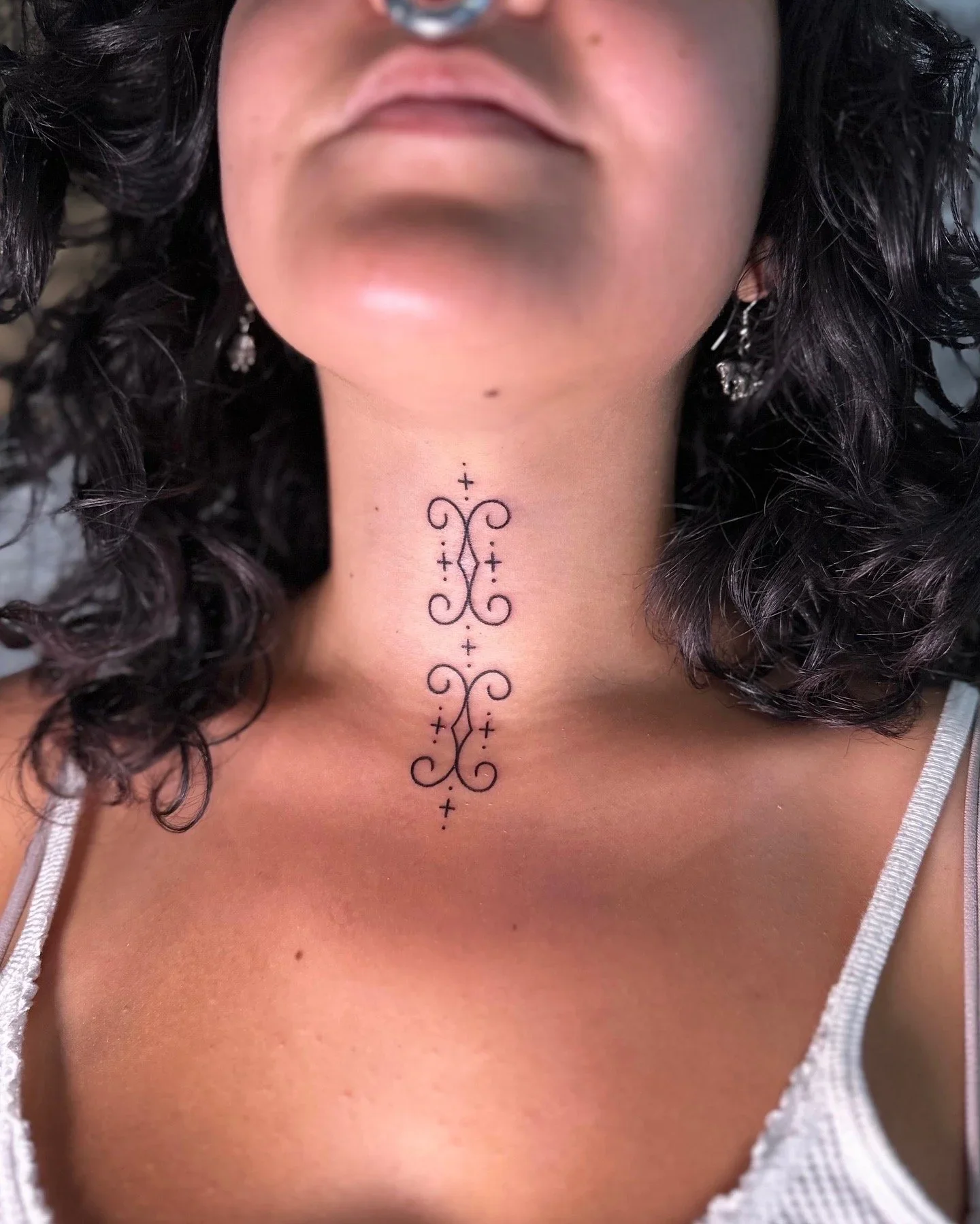 Close-up of a woman with curly dark hair wearing earrings, showcasing a black decorative tattoo on her neck. She's dressed in a white tank top.