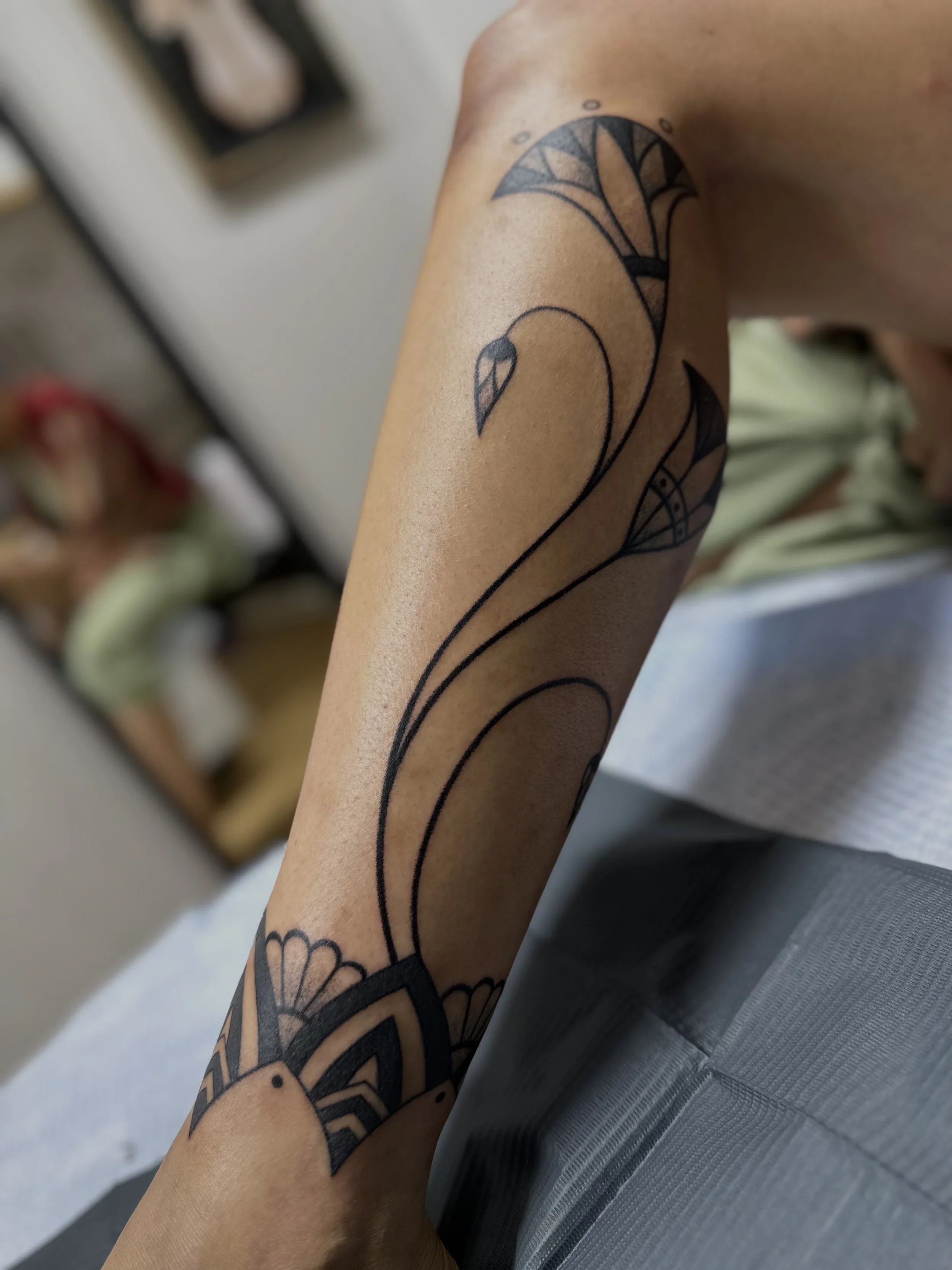 Close-up of a person's arm with a black and gray tattoo of a stylized flamingo on the forearm, with a blurred background.