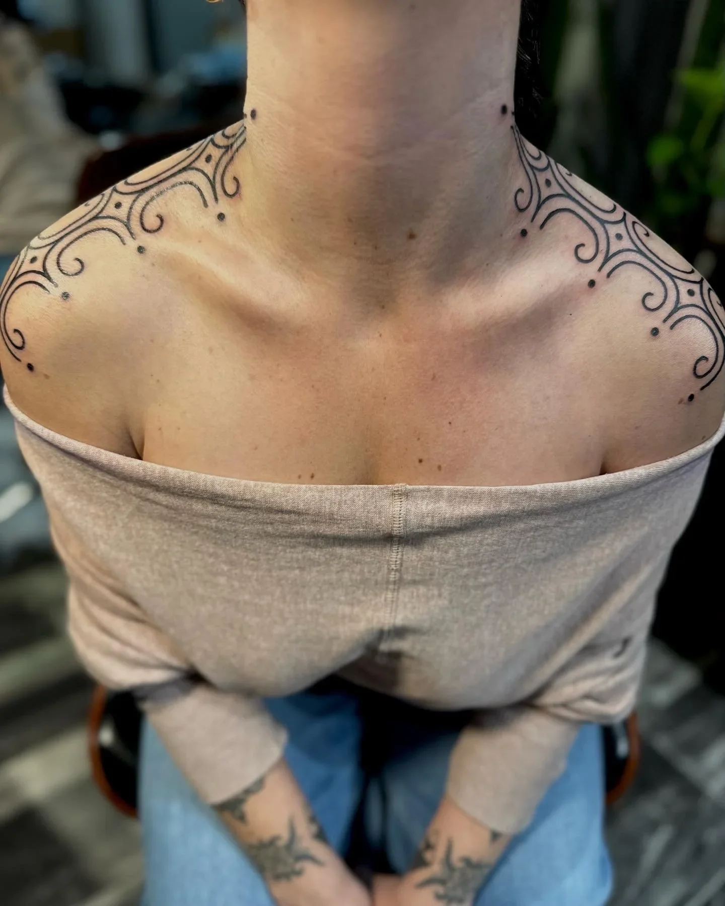 Close-up of a person's shoulders and neck with intricate black tattoo designs on the shoulders and upper chest, wearing a beige off-shoulder top.