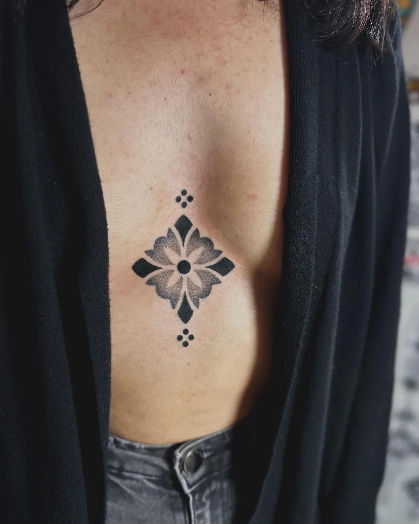 Close-up of a person with a tattoo on their chest, wearing a black top and jeans.