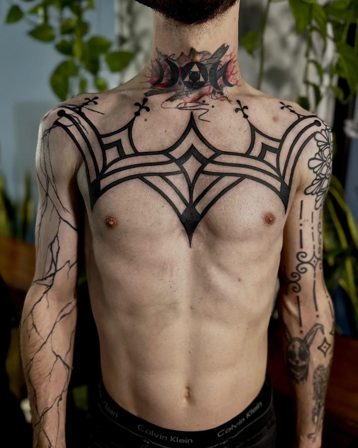 A shirtless man with numerous black tattoos, including a large symmetrical design across the chest, a tattoo on the neck with a triangle and eye, and various tattoos on the arms.