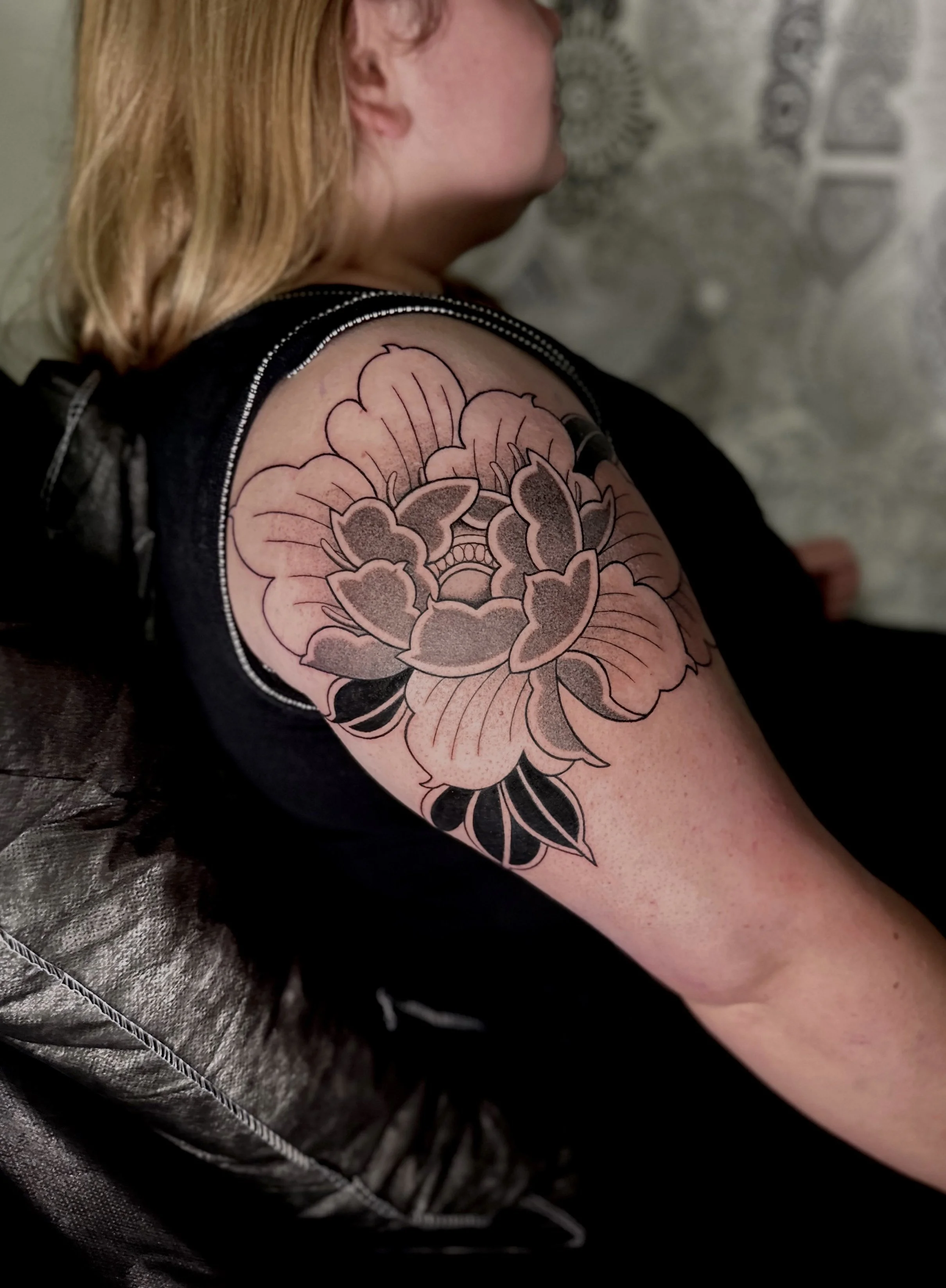 A person's upper arm with a large floral tattoo in black and pink ink. The tattoo features a detailed rose with leaves.