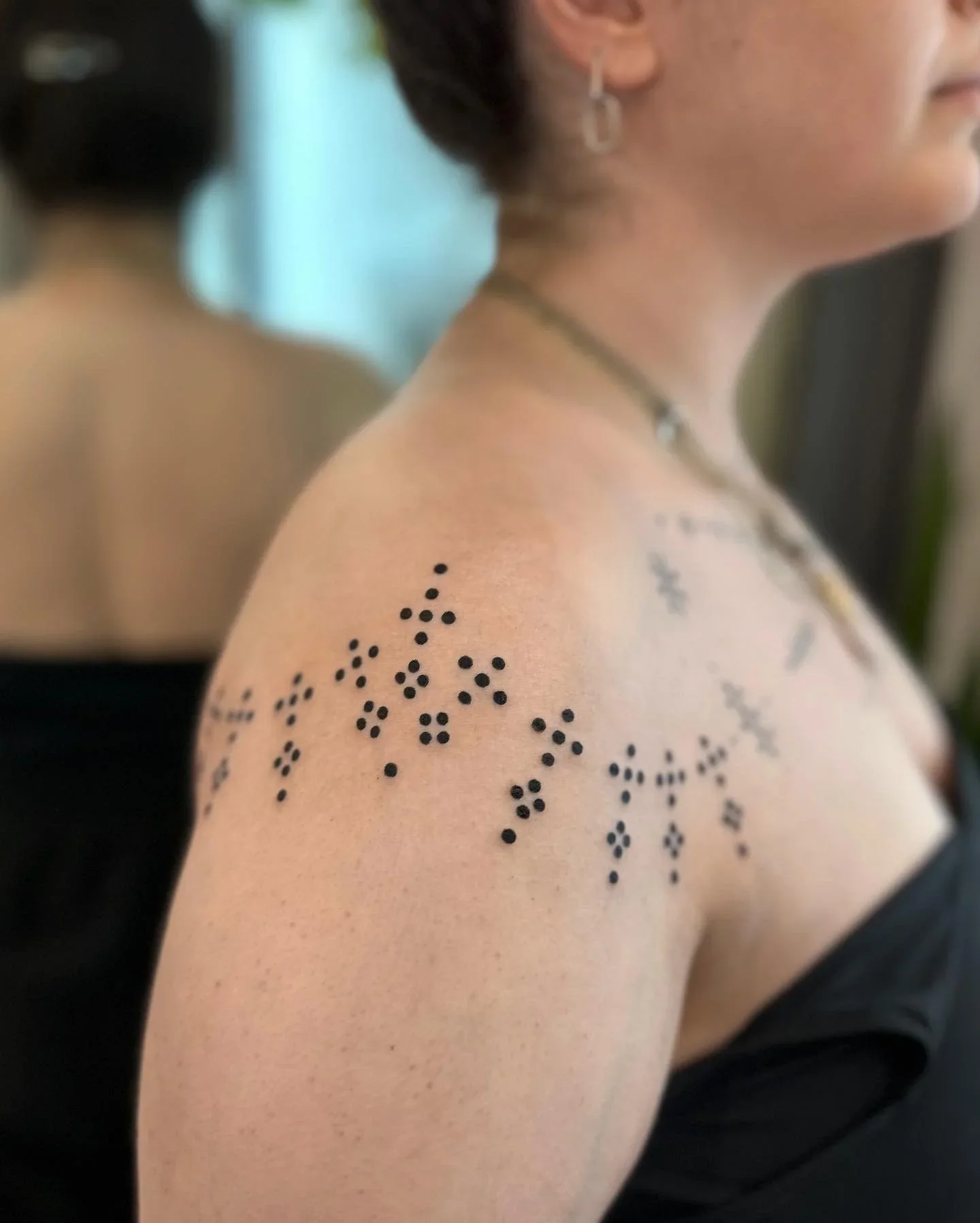 Close-up of a woman's shoulder and neck with a black floral tattoo and wearing a necklace, with blurred background.