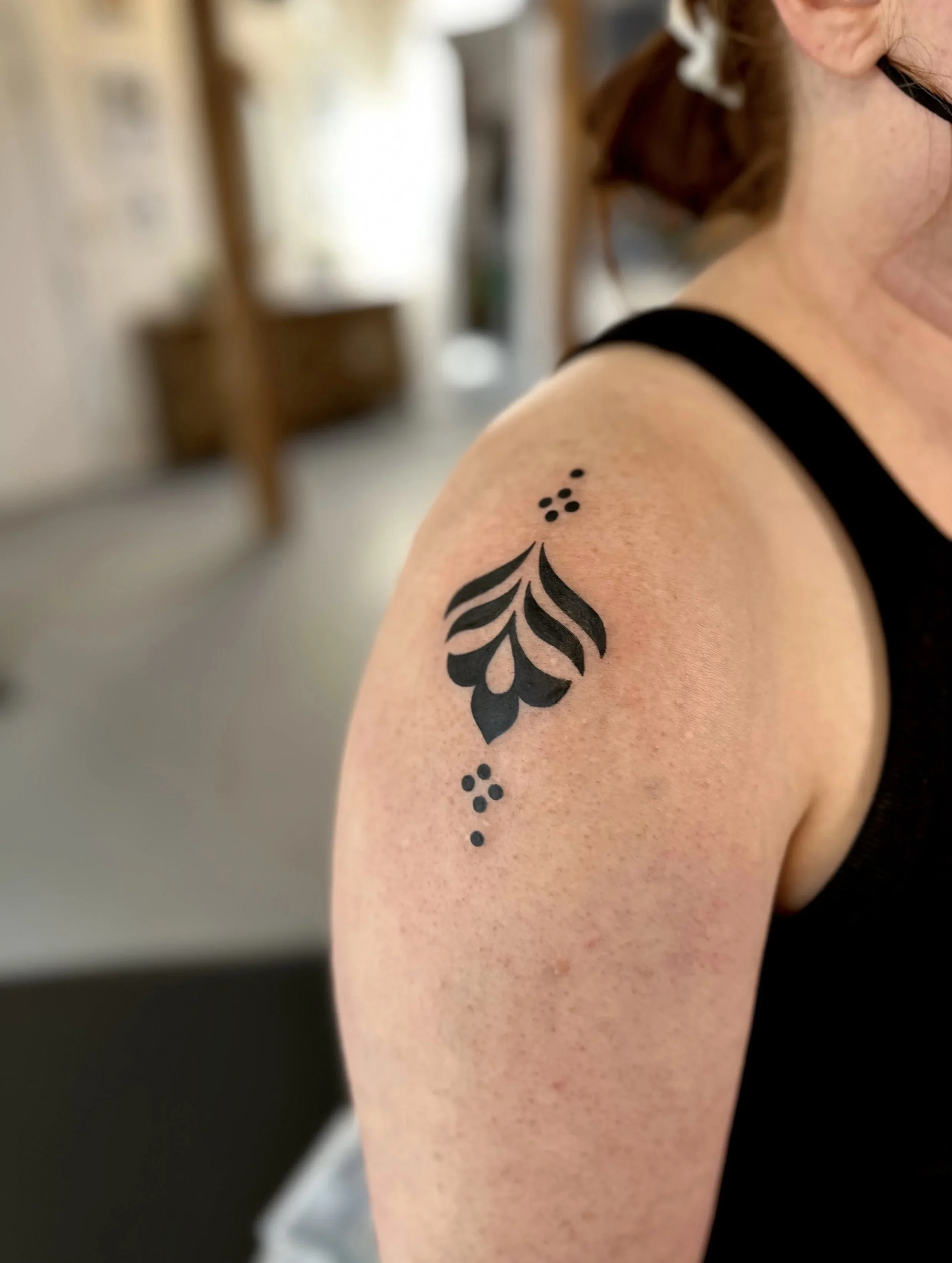 A person's shoulder with a black tattoo featuring a floral or leaf pattern and dotted accents.