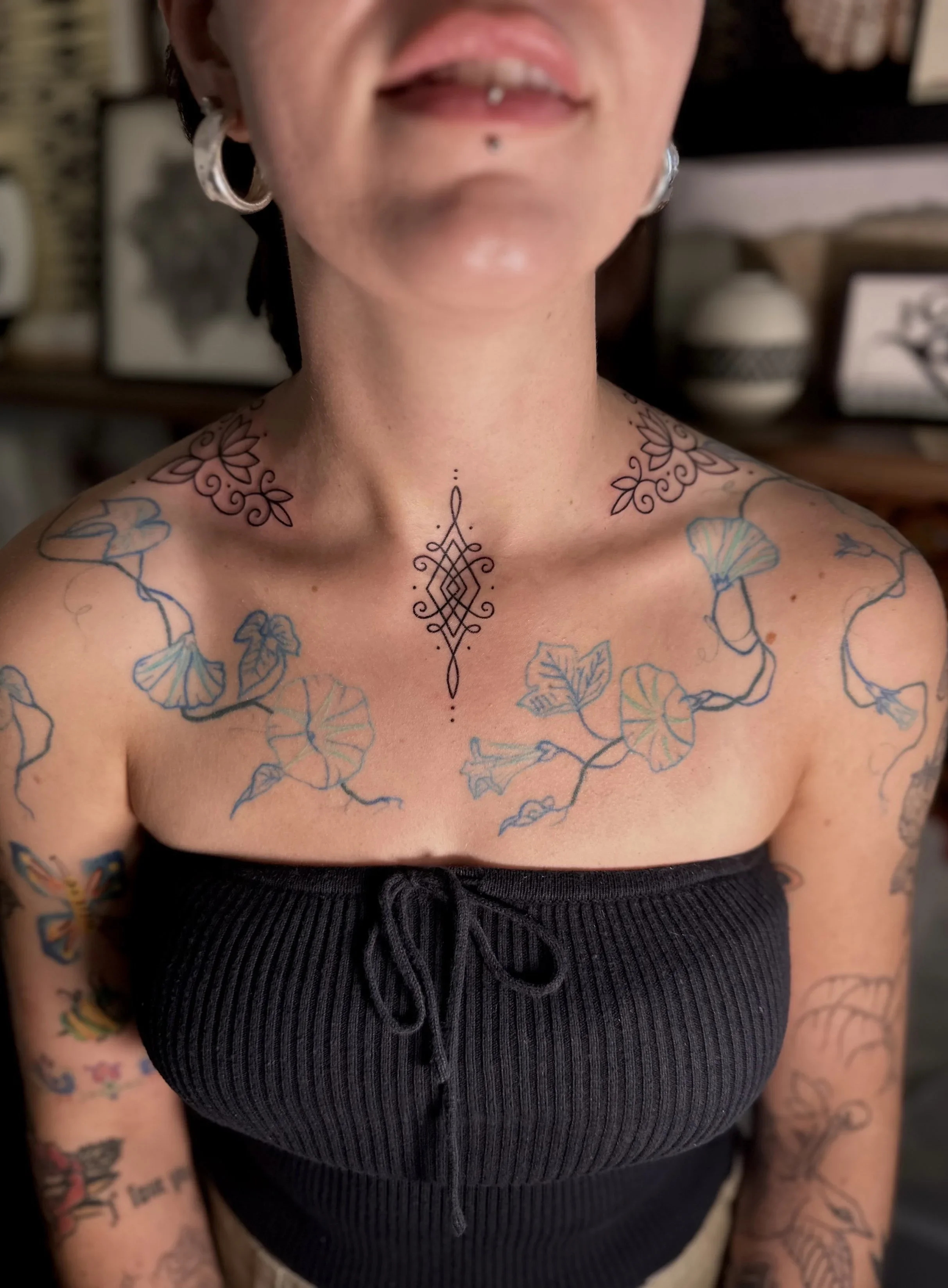 Close-up of a person's chest and shoulders, showing tattoos and wearing a black ribbed top, with a background of framed artwork and vases.