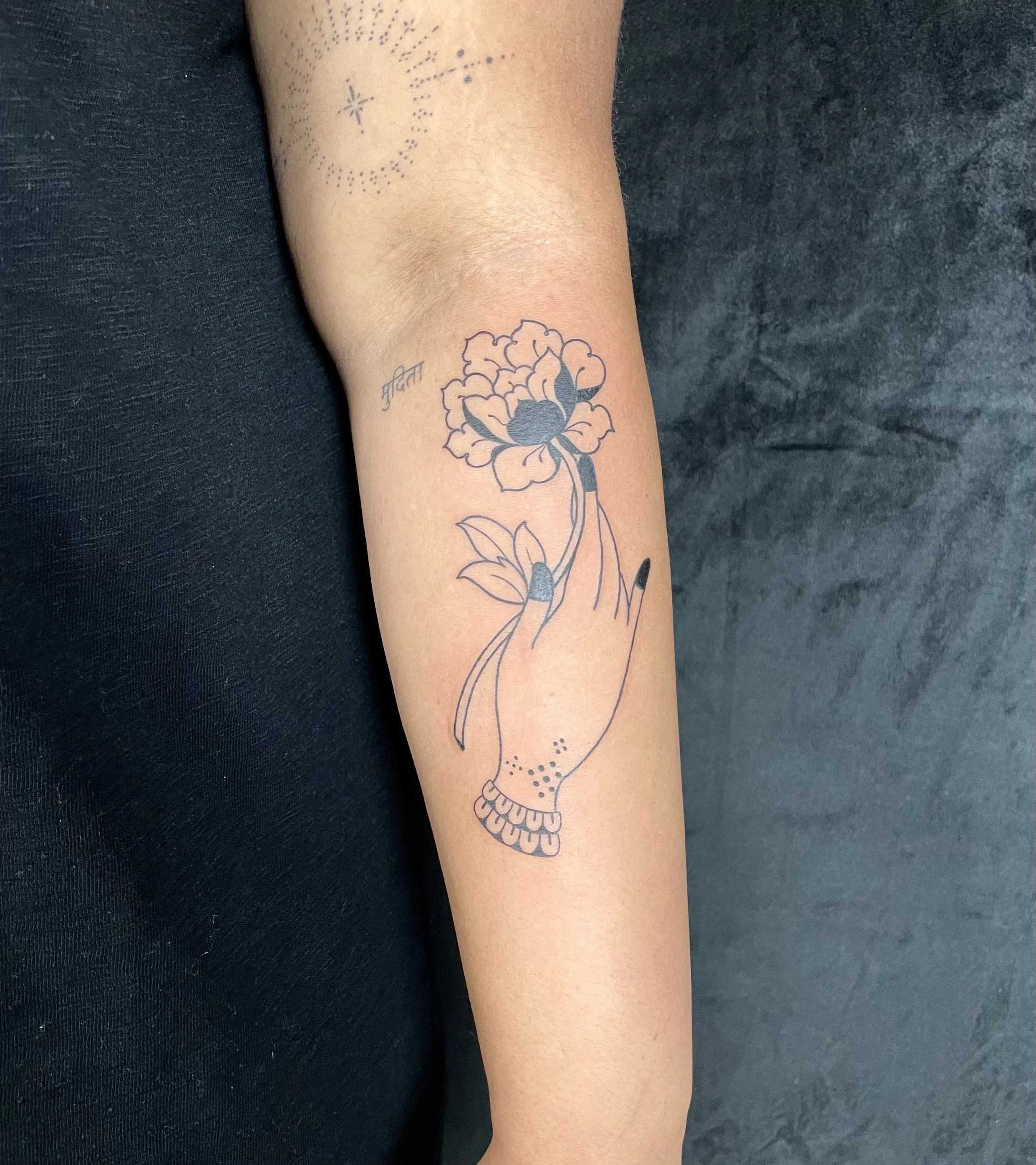 A person's forearm with a minimalistic tattoo of a flower with leaves and a stem, and small decorative elements including dots and a star shape, with a small handwritten word in a different language near the flower.