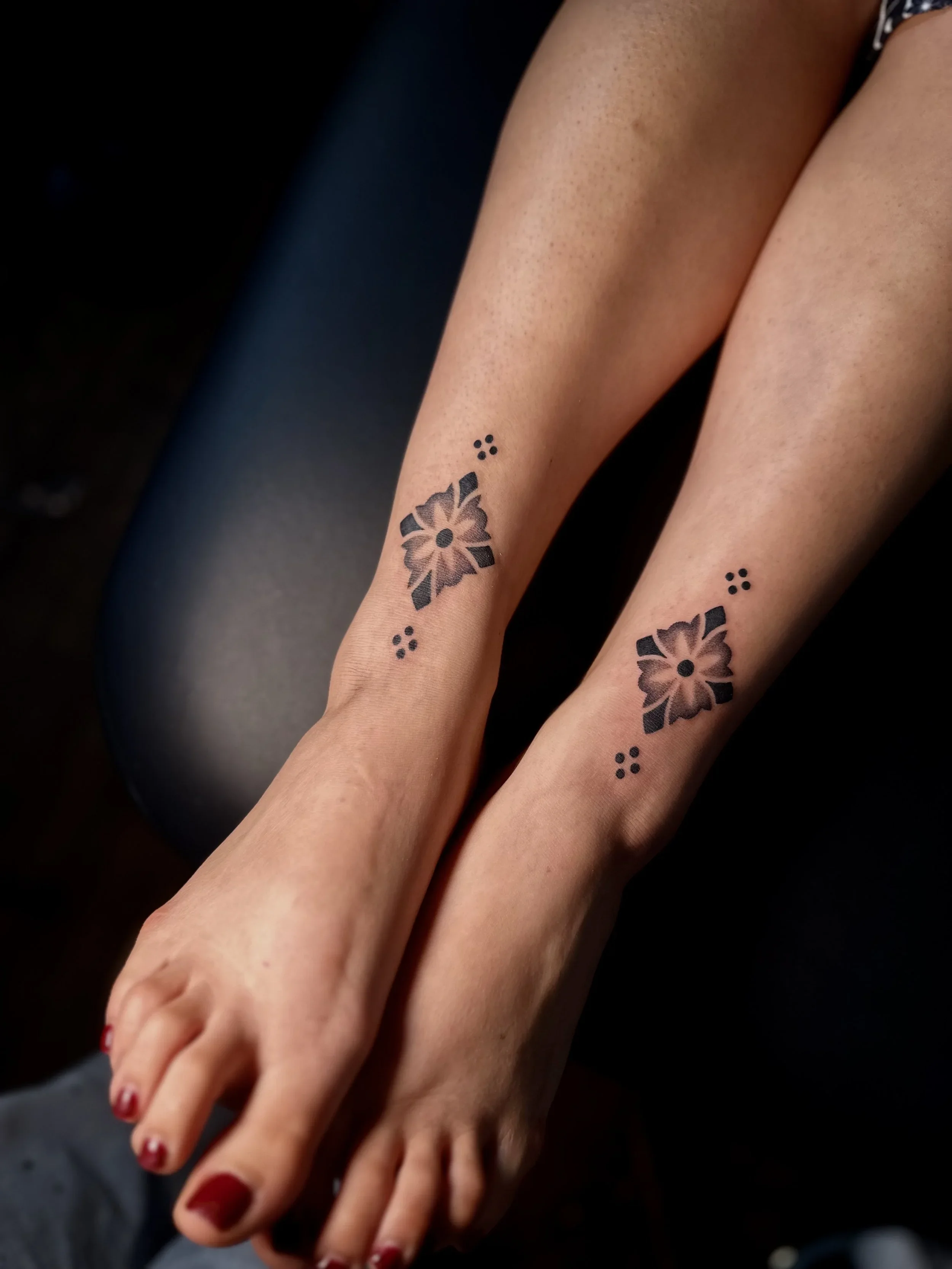 Close-up of two legs with black floral tattoos and red nail polish on toes, resting on a dark surface.