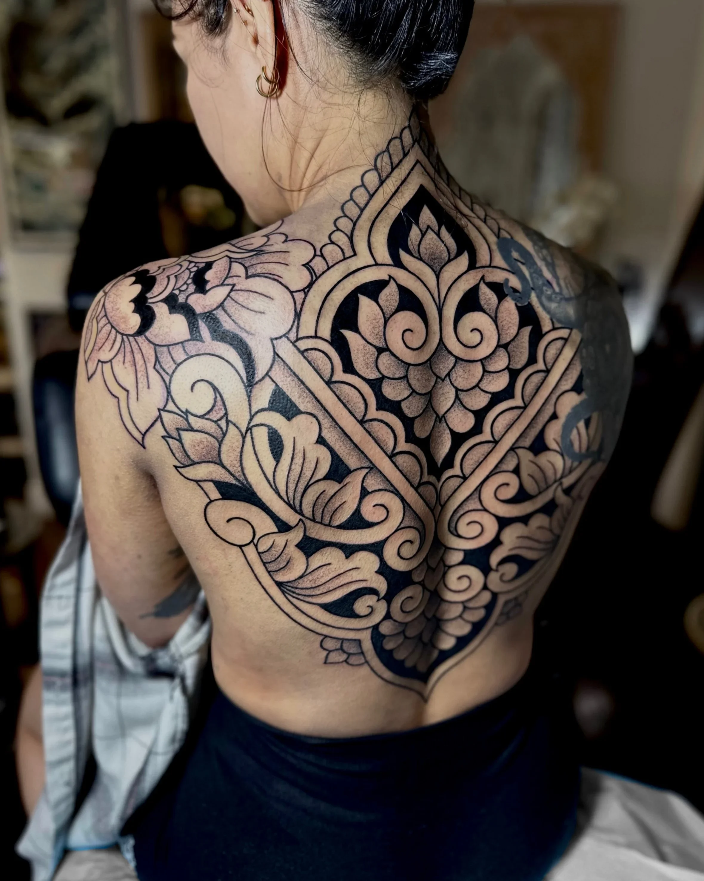 Back view of a person's shoulder and upper back with an intricate black and gray tattoo design. The tattoo features floral and geometric patterns that extend from the shoulder to the middle of the back. The person has dark hair pulled back and is wea