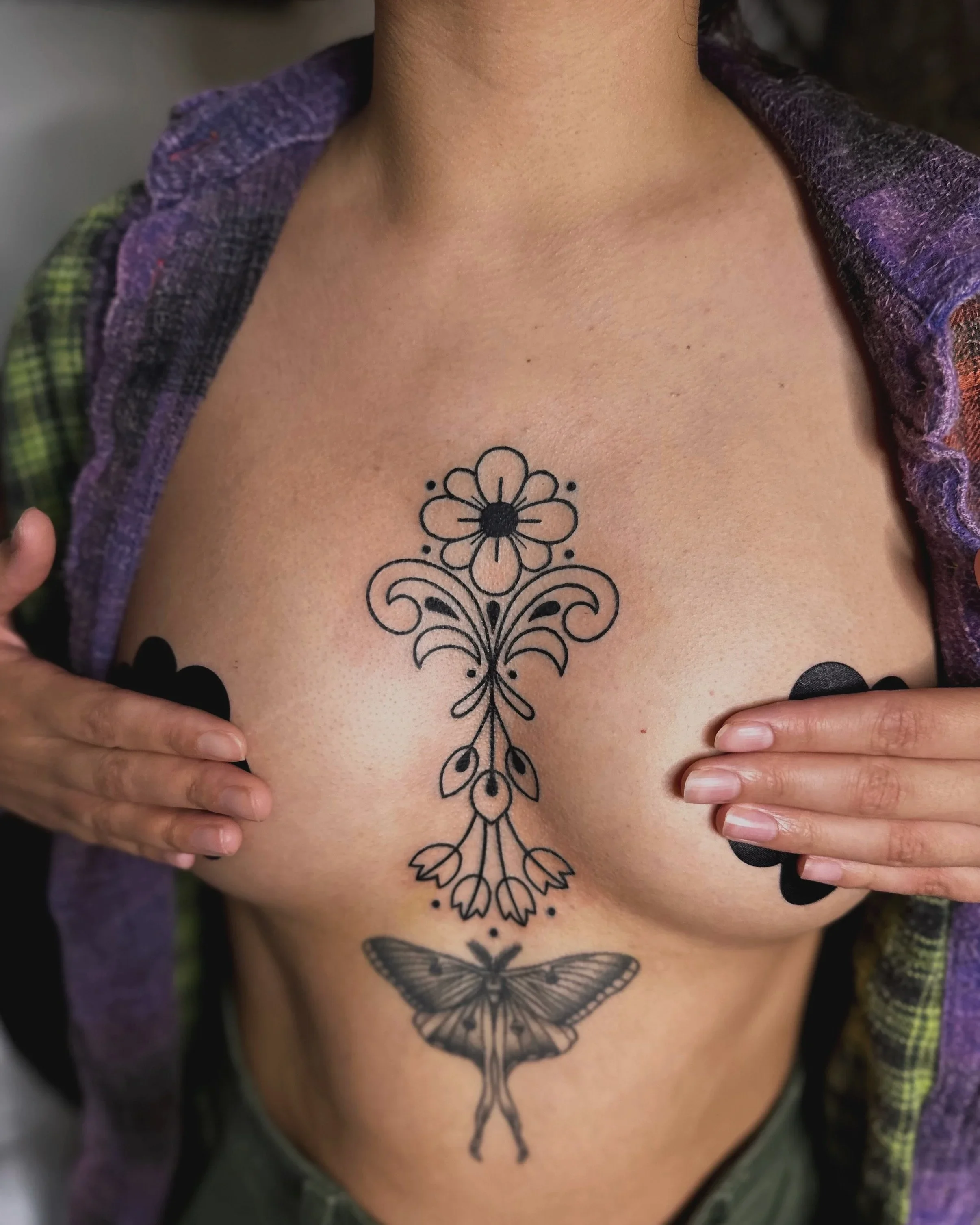 Close-up of a person's chest with a black butterfly tattoo, with hands holding the sides of their torso, revealing the tattoo. The person is wearing a purple garment with a textured, woven pattern.